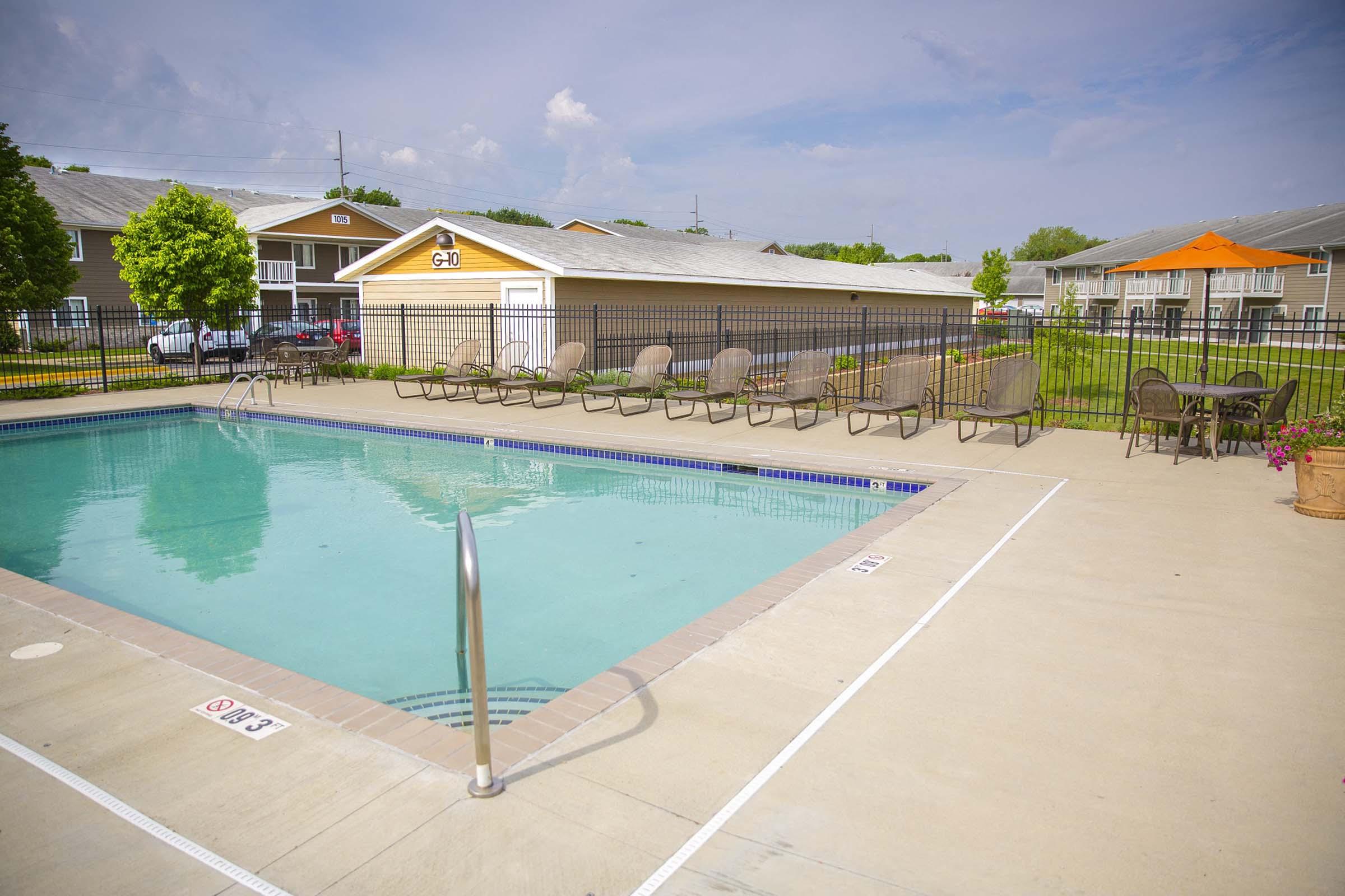 A sunny outdoor swimming pool surrounded by lounge chairs, with a fence and grassy area nearby. A small building is visible in the background, and residential units can be seen beyond the pool area. The sky is partly cloudy, indicating a pleasant weather day.