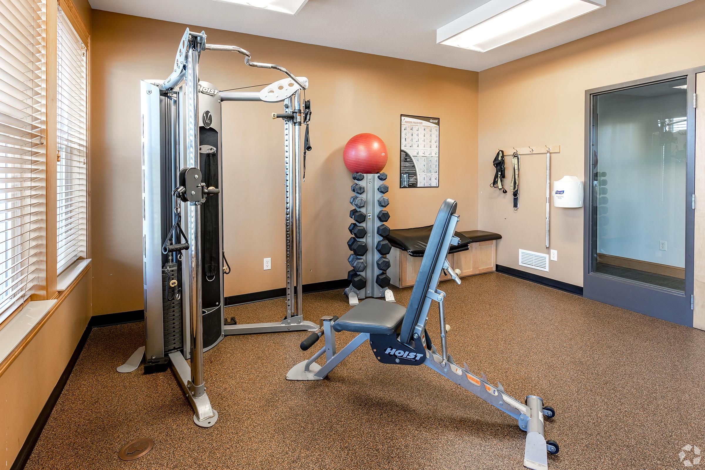 A small gym interior featuring a cable machine, a stability ball, a weight rack, and a flat bench. The room has windows allowing natural light, with a beige and brown color scheme. A calendar and a wall-mounted dispenser are visible in the background.