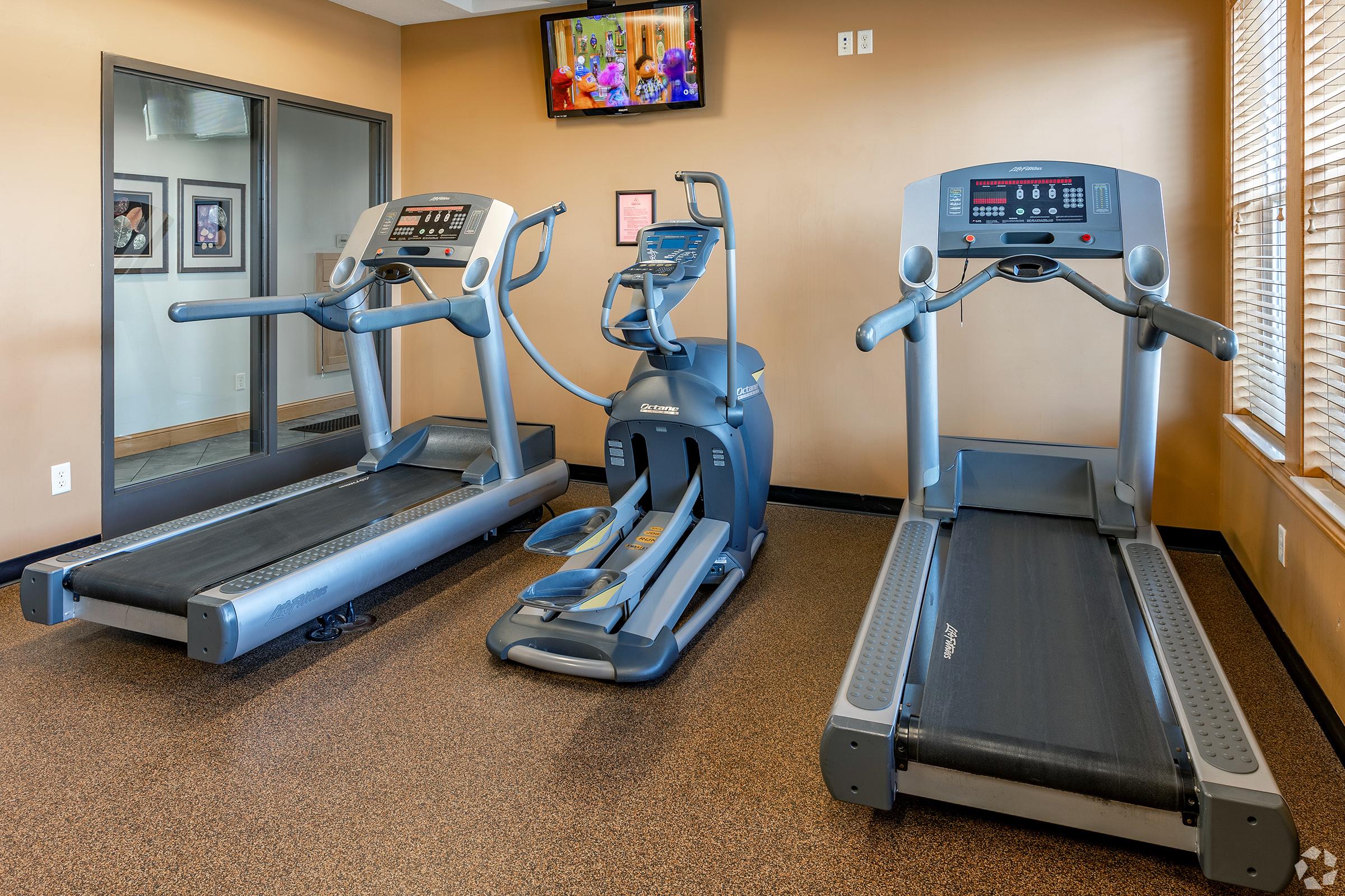 A fitness room featuring three exercise machines: two treadmills side by side and one elliptical machine in the center. The walls are painted a warm brown, and there is a television mounted on the wall showing a colorful display. Natural light filters in through a window with blinds.