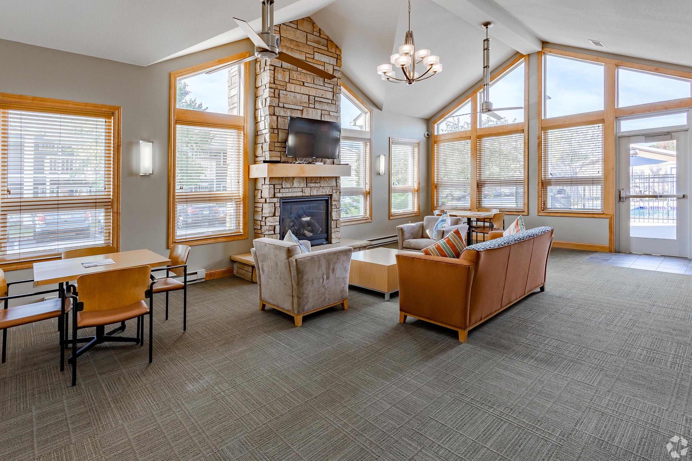 Cozy lounge area featuring large windows with natural light, a stone fireplace, a television mounted on the wall, and comfortable seating arrangements. There are two tables set with chairs, and a door leading to an outdoor area, creating an inviting atmosphere for relaxation and gatherings.
