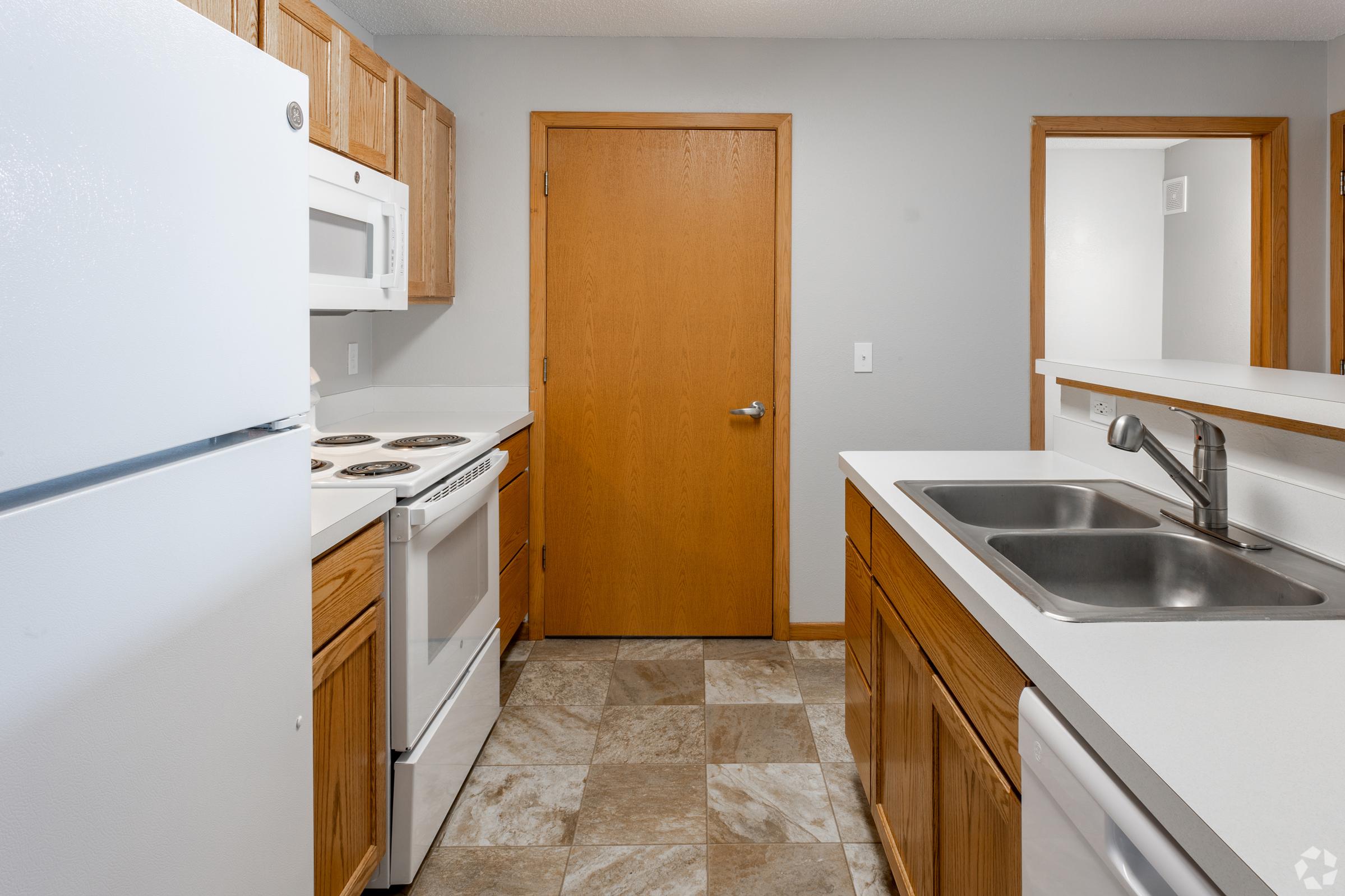 A modern kitchen featuring light wood cabinets, a white refrigerator, an oven with a stovetop, and a microwave. The countertop is white with a double sink, and the floor has gray and beige tiles. A wooden door leads to another room in the background.