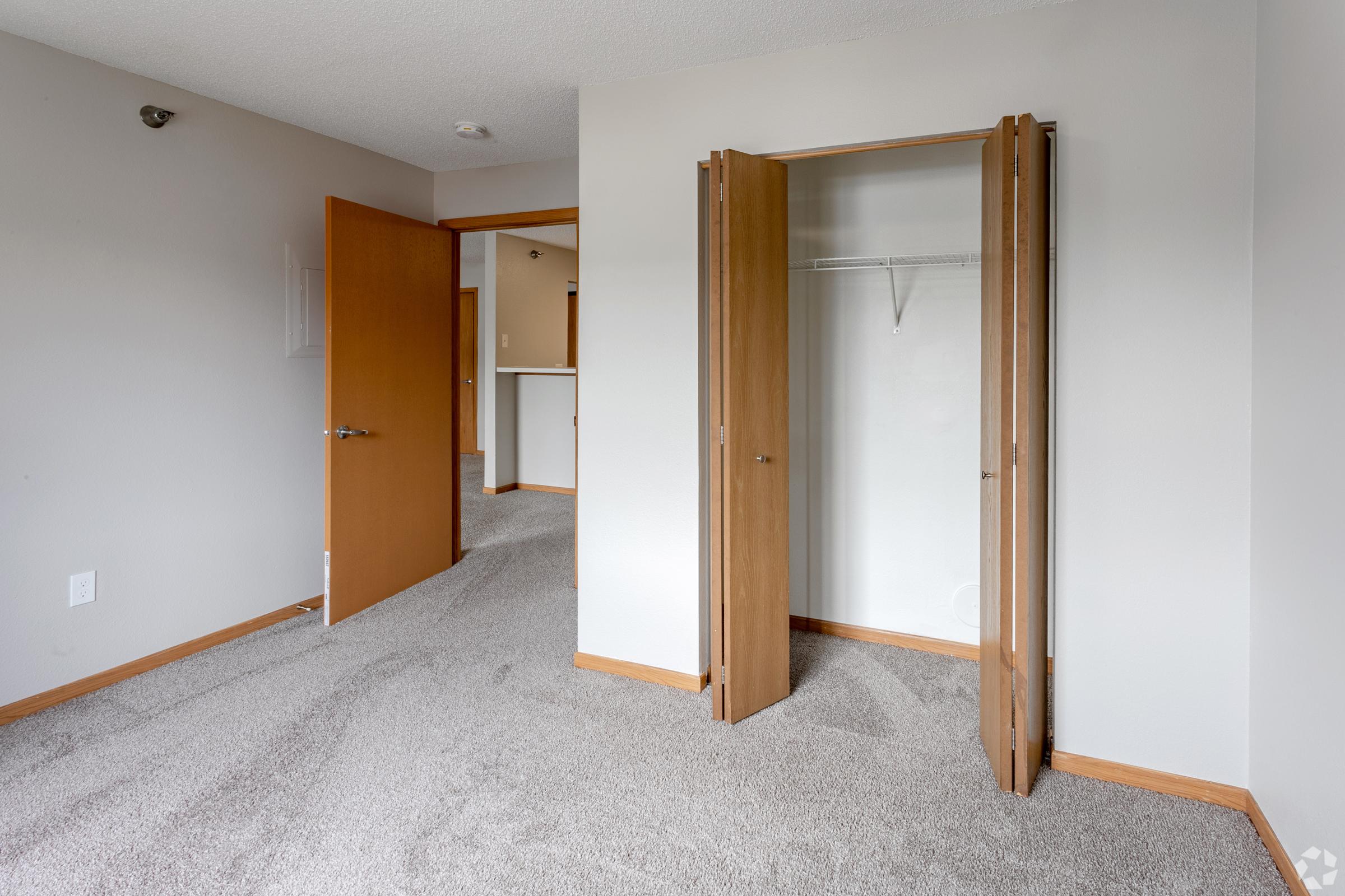 Interior view of a vacant room featuring light-colored walls and carpeting. There are two doors visible: one leading to another room and the other to a closet with sliding doors. The space is well-lit, with neutral tones creating an inviting atmosphere.