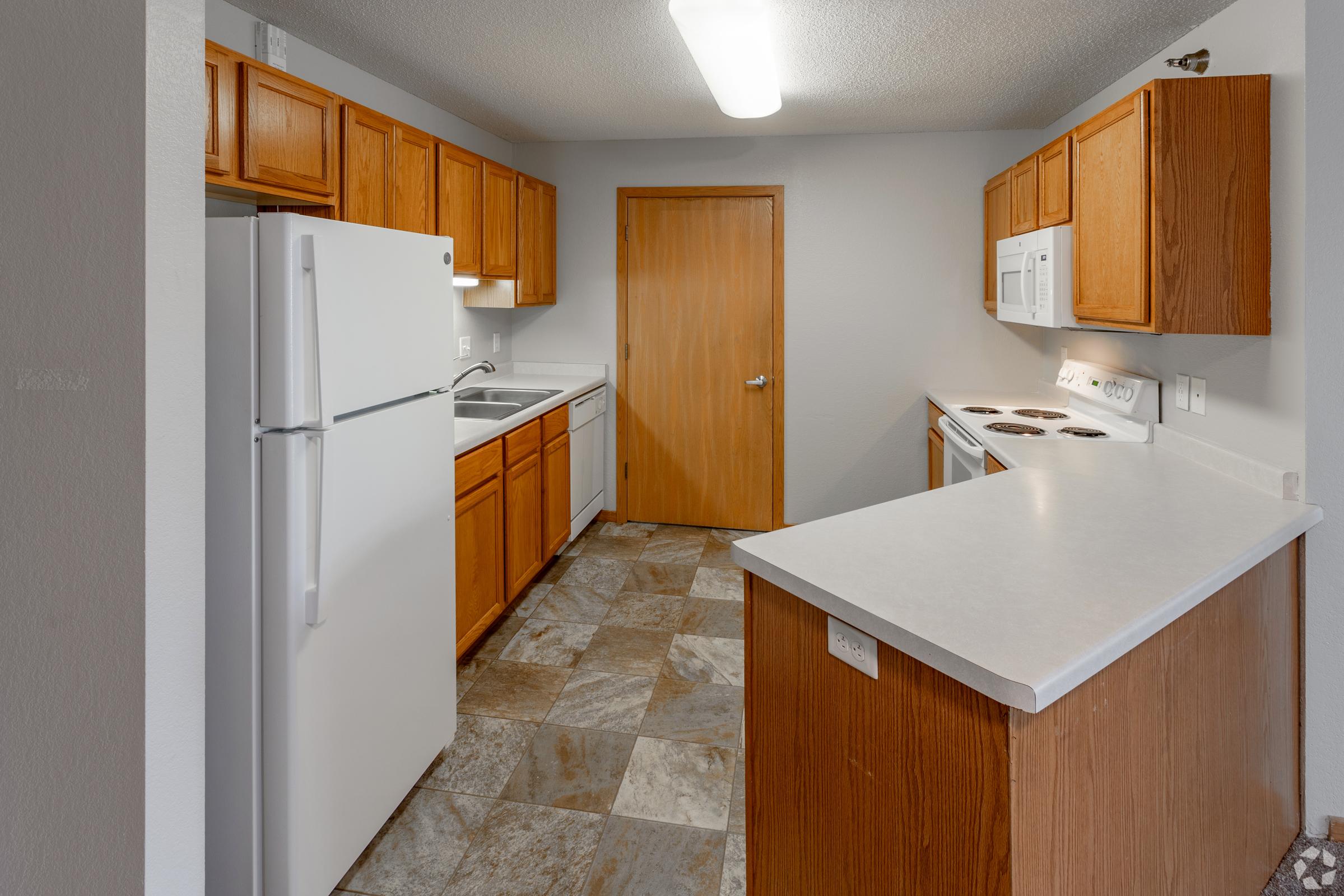 A modern kitchen featuring wooden cabinets, a white refrigerator, stove, and microwave. The space has a light-colored countertop and tile flooring, with a door leading to another room. Natural light illuminates the area from a ceiling fixture.