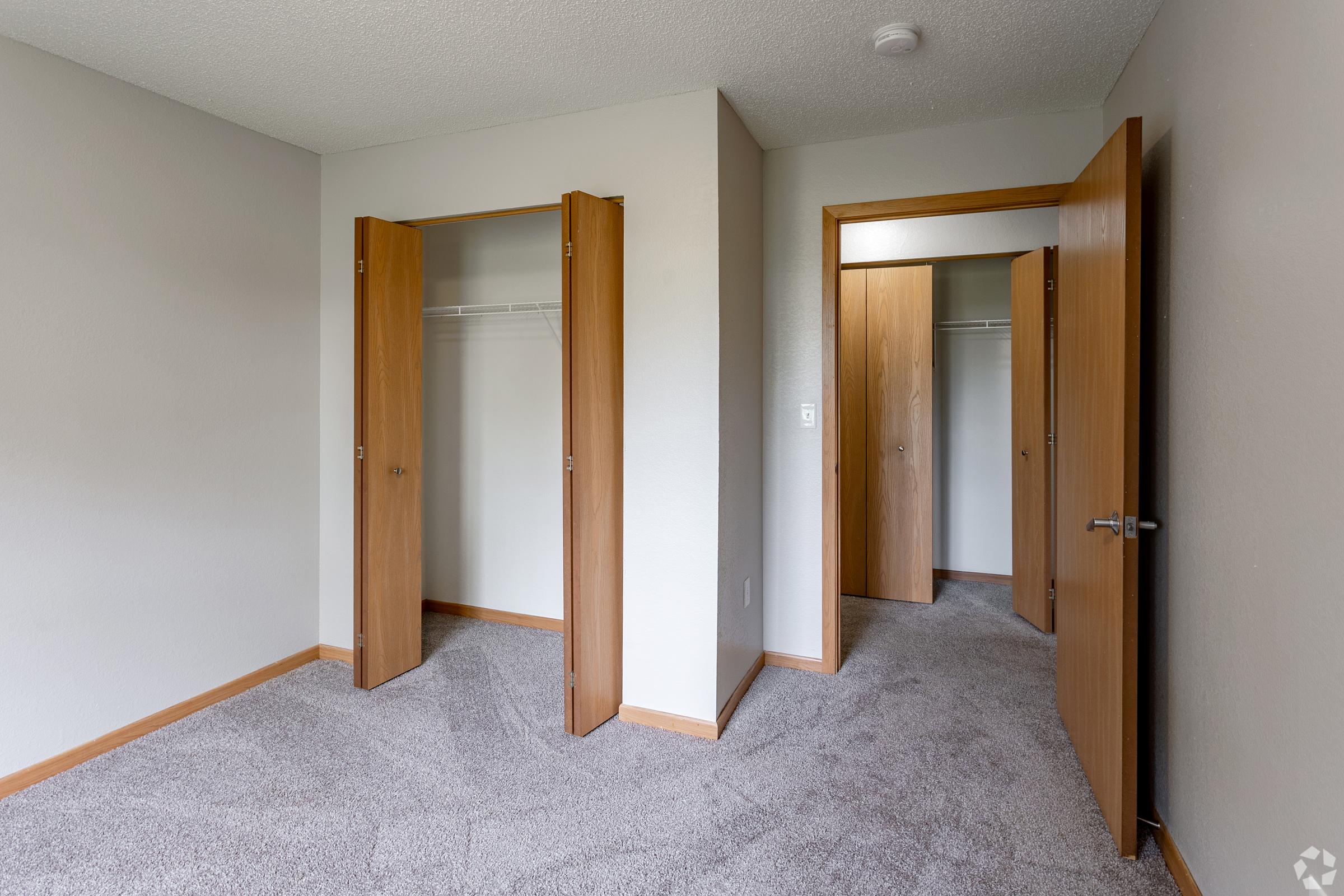 A lightly furnished room with beige carpet. Two wooden doors lead to closets on the left and right walls. The walls are painted in a neutral color, and there is a ceiling light fixture. The room appears bright and empty, suitable for various uses like a bedroom or office.