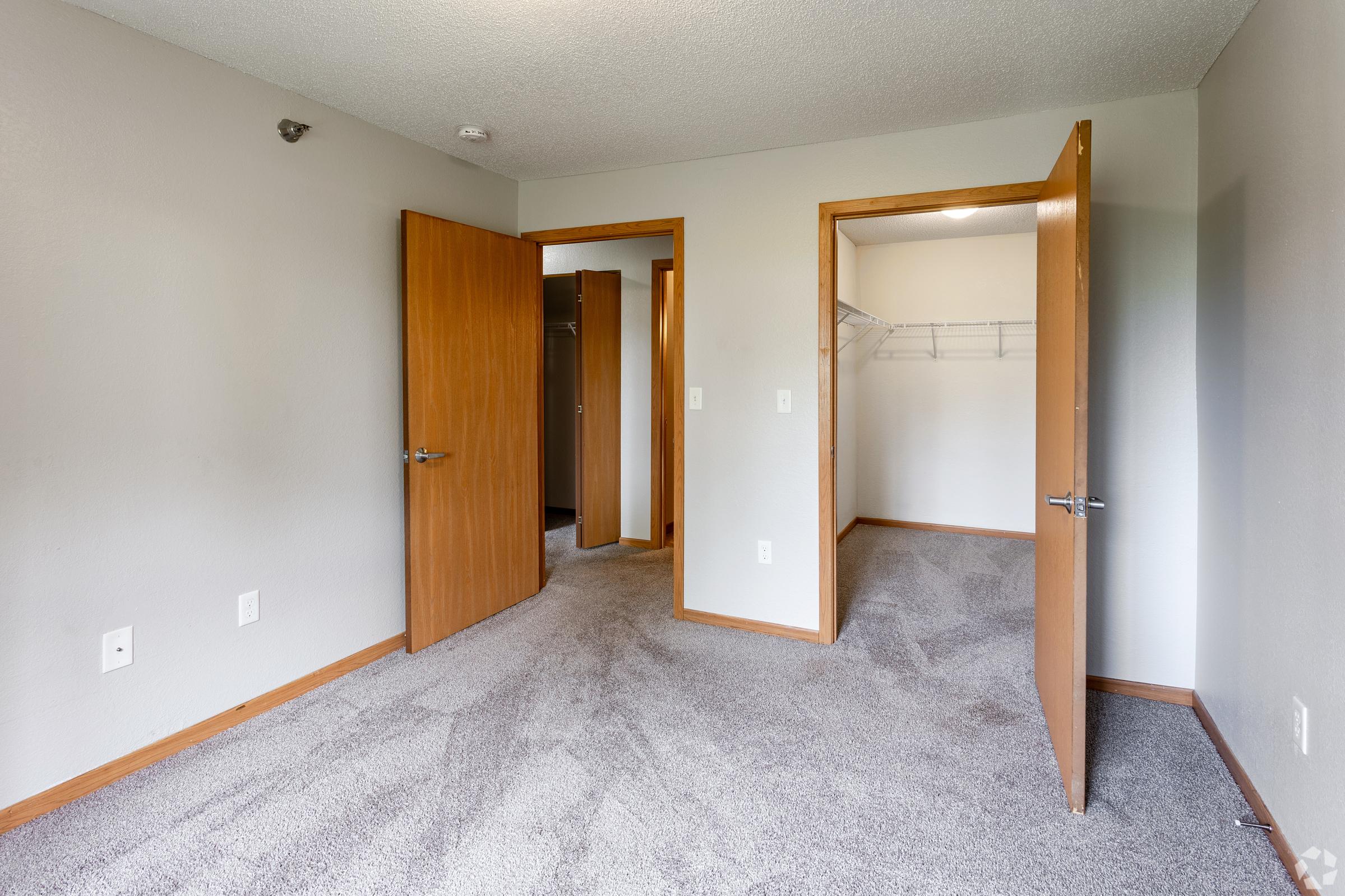 A well-lit, empty room with light gray carpet and neutral walls. Two open wooden doors lead to a closet on the right and another room on the left. The space is uncluttered, showcasing clean lines and simple design, suitable for various uses such as a bedroom or office. Natural light illuminates the area.