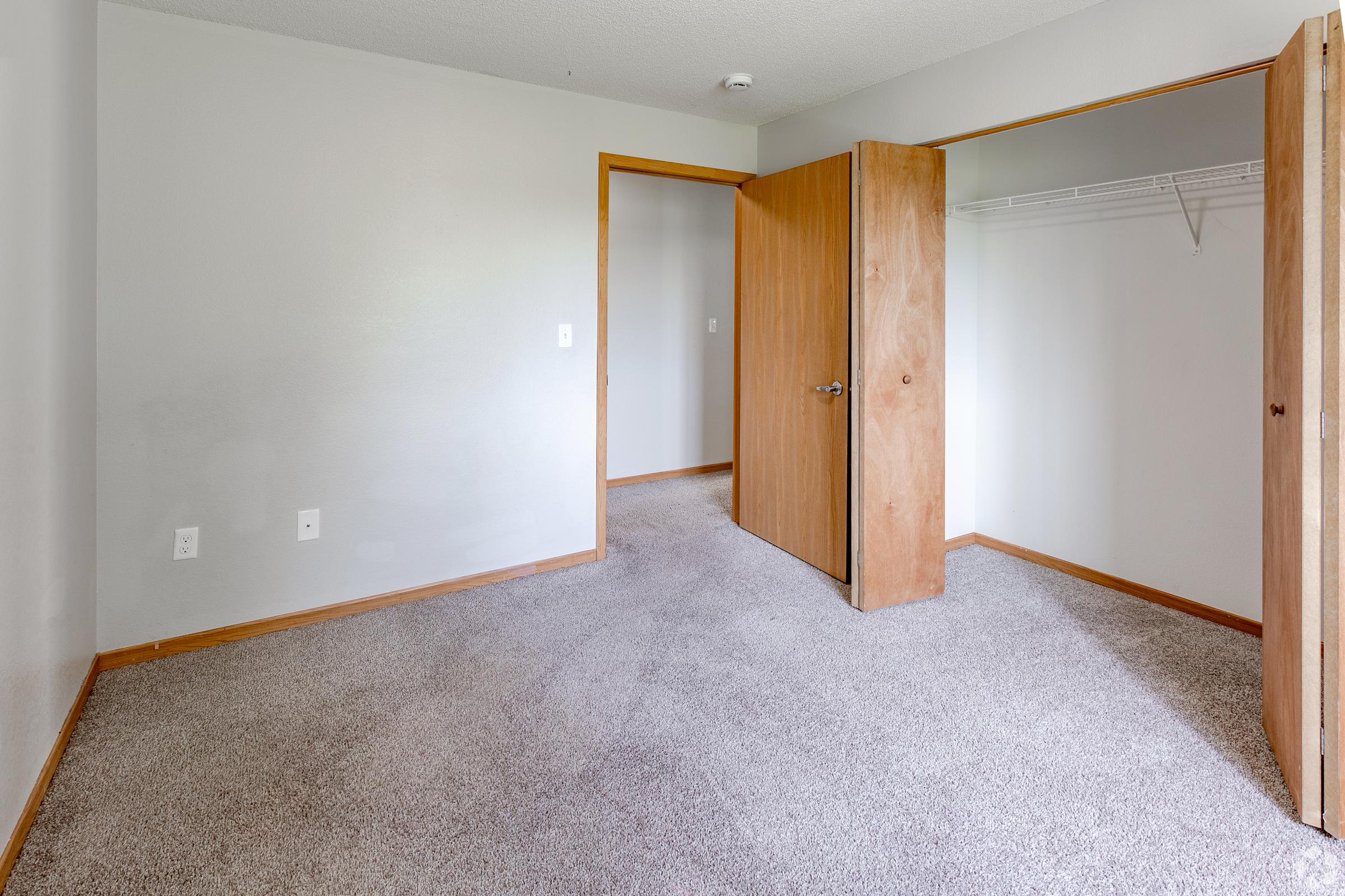 A spacious, empty room with beige walls and a gray carpet. There's a wooden door partially open leading to a closet and another door leading to a hallway. Natural light illuminates the space, creating a clean and inviting atmosphere.