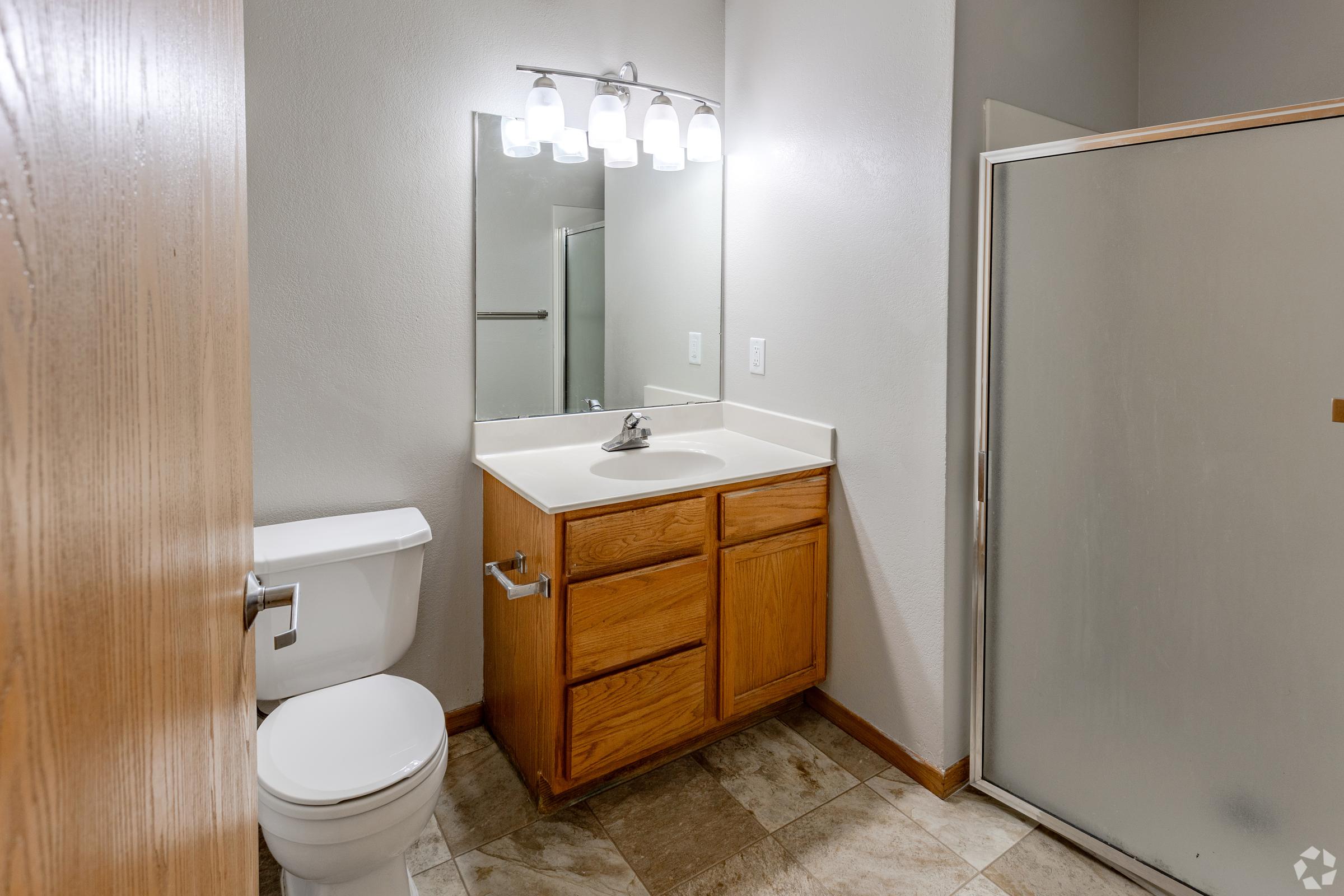 A clean and modern bathroom featuring a wooden cabinet vanity with a sink, a toilet, and a glass shower enclosure. The walls are painted light gray, and there is overhead lighting offering bright illumination. The floor is tiled with large rectangular tiles.