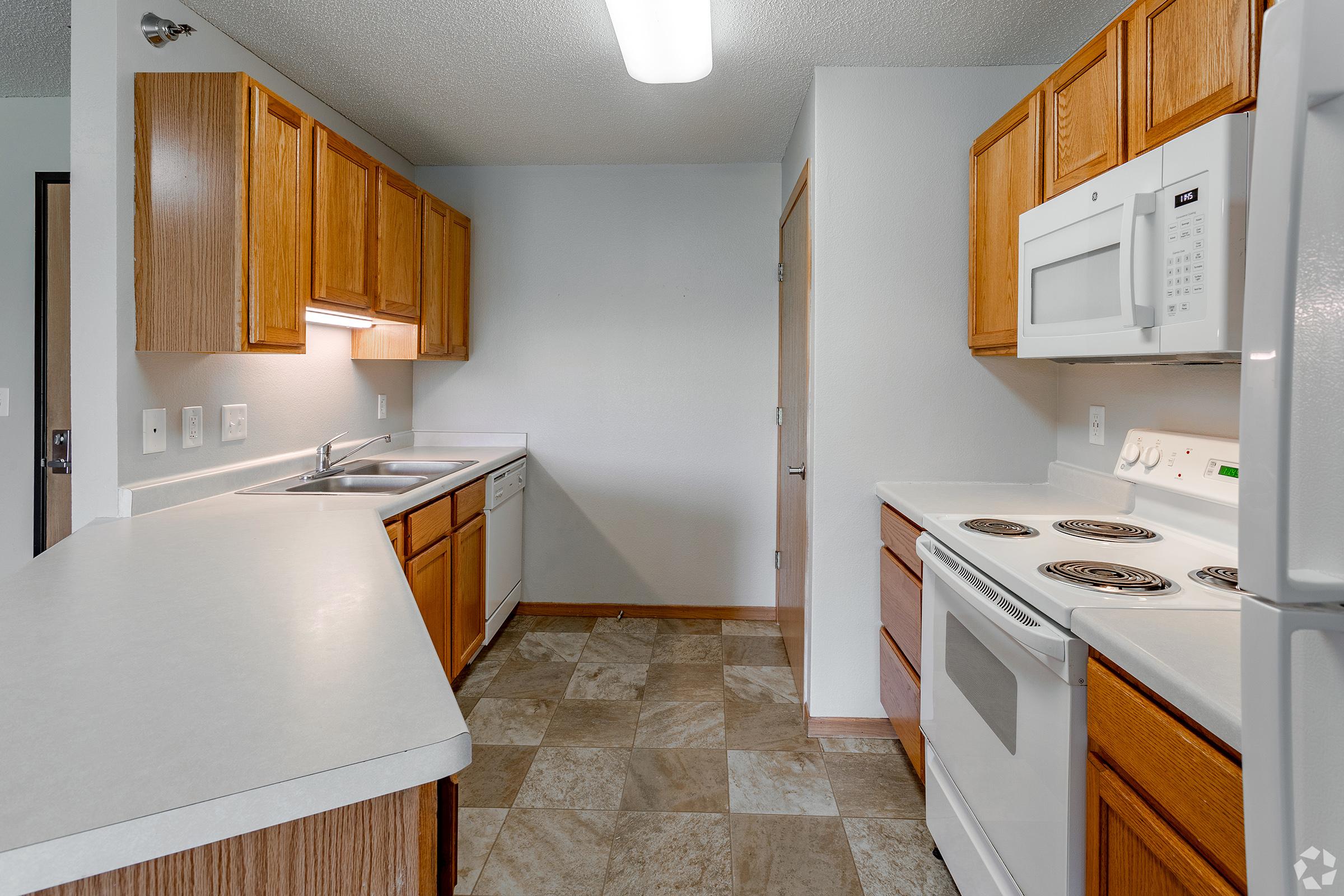 A small, modern kitchen featuring wooden cabinetry, white countertops, and a tile floor. The kitchen includes a sink, stove, microwave, and oven, with ample natural light coming from a ceiling fixture. The layout is U-shaped, optimizing space for cooking and meal preparation.