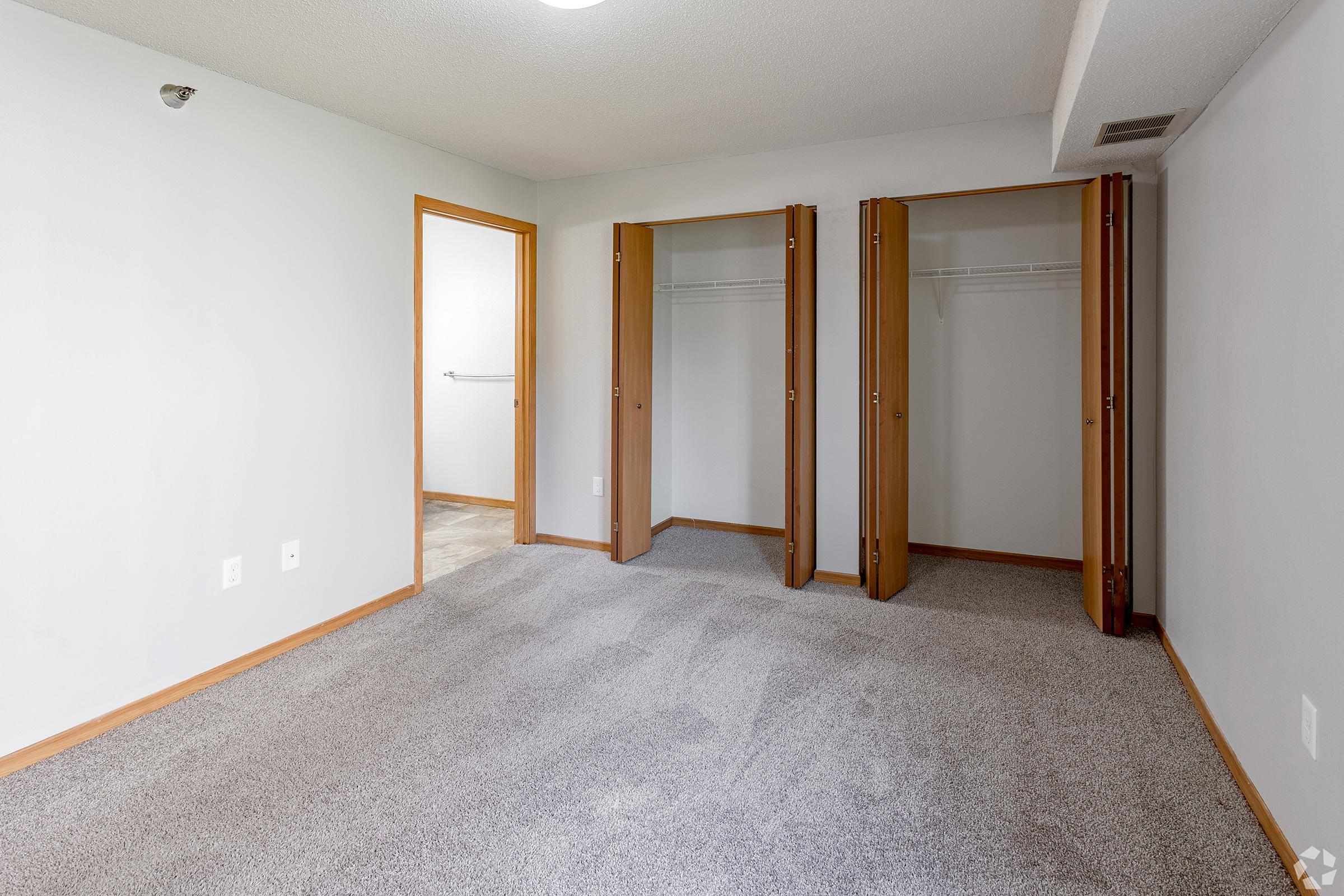 A spacious, empty room with light gray walls and carpet. Two open closets with wooden doors are on the right side, showcasing hanging space. A doorway on the left leads to another room, and the ceiling features a light fixture. The overall atmosphere is bright and clean.
