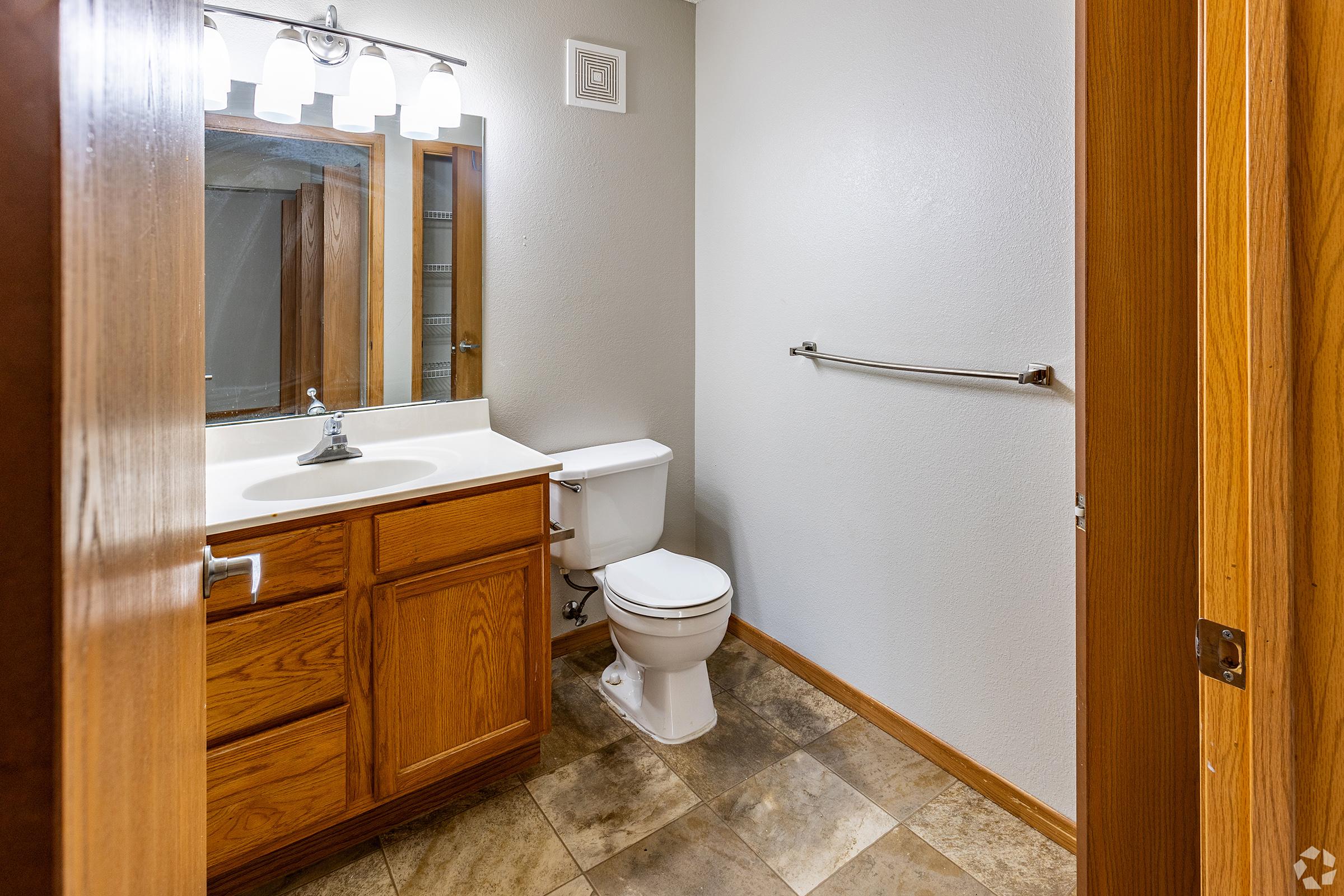 A clean bathroom featuring a white toilet, a wooden vanity with a sink, and a mirror above it. The walls are painted light gray, and the floor is tiled with various shades of brown. There is a towel rack mounted on the wall and wooden doors framing the entrance.