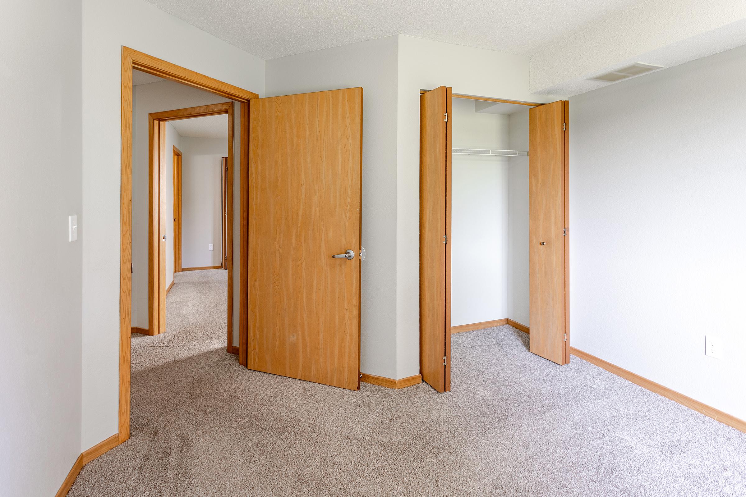 A well-lit room with light gray walls and carpeted flooring. The room features two wooden doors leading to adjacent areas, one of which opens to a closet. Natural light enters through the open doors, creating a spacious feel. The decor is simple and modern, emphasizing minimalism.