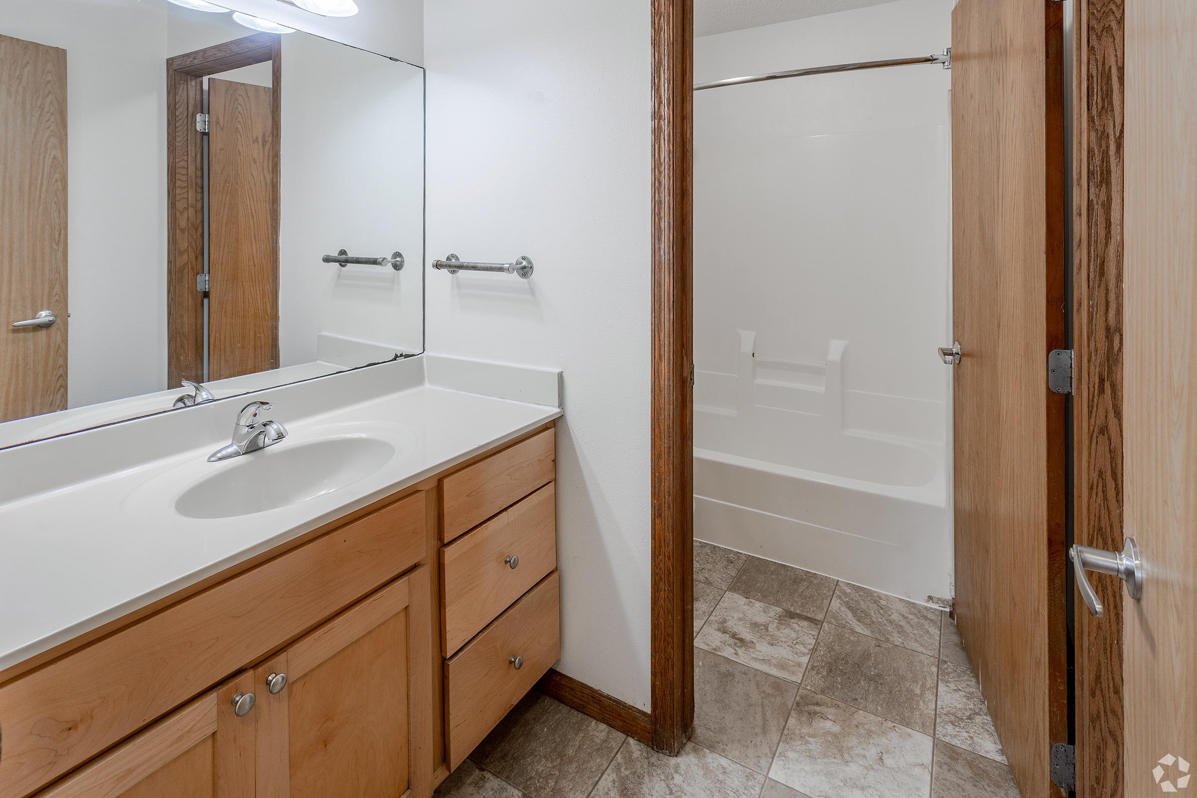 A clean and modern bathroom featuring a beige countertop with a sink, large mirror, wooden cabinetry, and a shower-tub combo. The floor is tiled with gray stone-like patterns, and the room has wooden door frames leading to an interior space. Bright overhead lighting enhances the spacious feel.