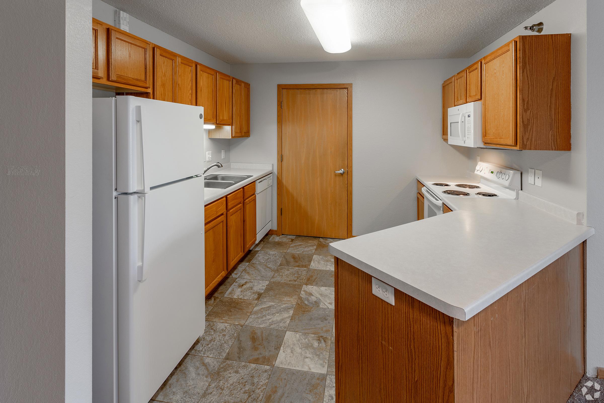 A view of a modern kitchen featuring wooden cabinets, a white refrigerator, a white microwave, and an electric stove. The countertops are light-colored, and the floor is covered with multi-toned tiles. A wooden door leads to another room, enhancing the spaciousness of the area.