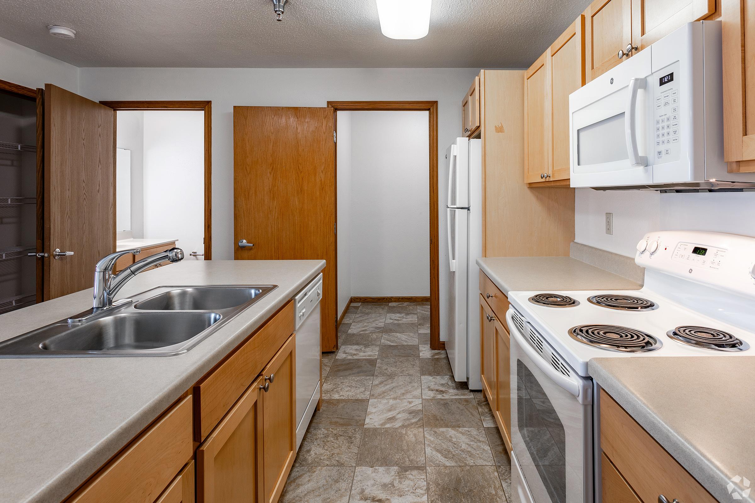 A modern kitchen featuring light wood cabinetry, stainless steel appliances including a microwave and dishwasher, dual sinks, and a smooth-top stove. The space has a neutral-colored tiled floor, ample counter space, and multiple doorways leading to other areas. Good lighting from a ceiling fixture enhances the room.