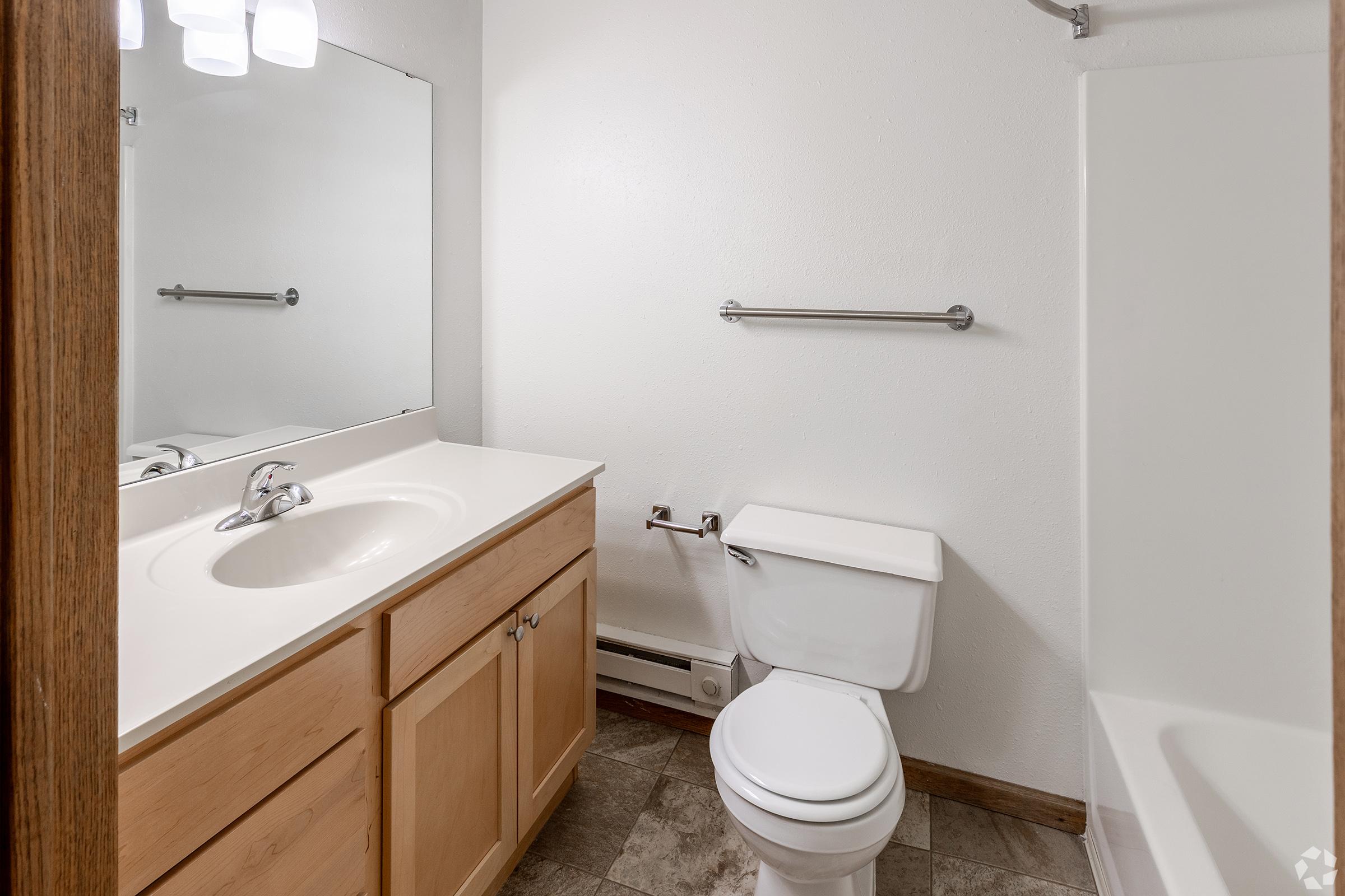 A clean and simple bathroom featuring a light-colored countertop with a sink, a mirror above, a wooden cabinet below, and a white toilet. There's a shower and bathtub combination on one side, with a towel rack mounted on the wall, and neutral-colored flooring.