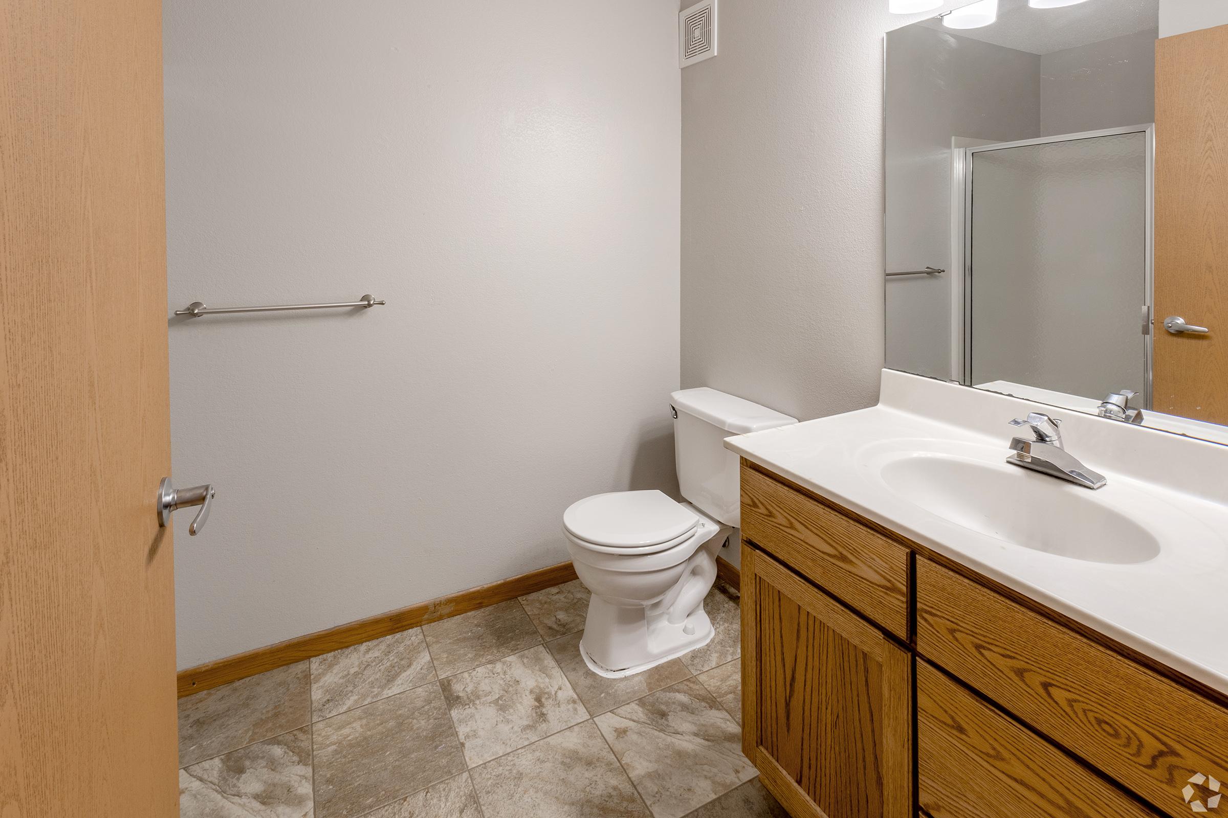 A clean, modern bathroom featuring a white toilet, a light-colored countertop with a silver faucet, and wooden cabinetry. The walls are painted in a neutral gray, and the floor is tiled with a light stone pattern. A shower stall is visible in the corner with a glass door.