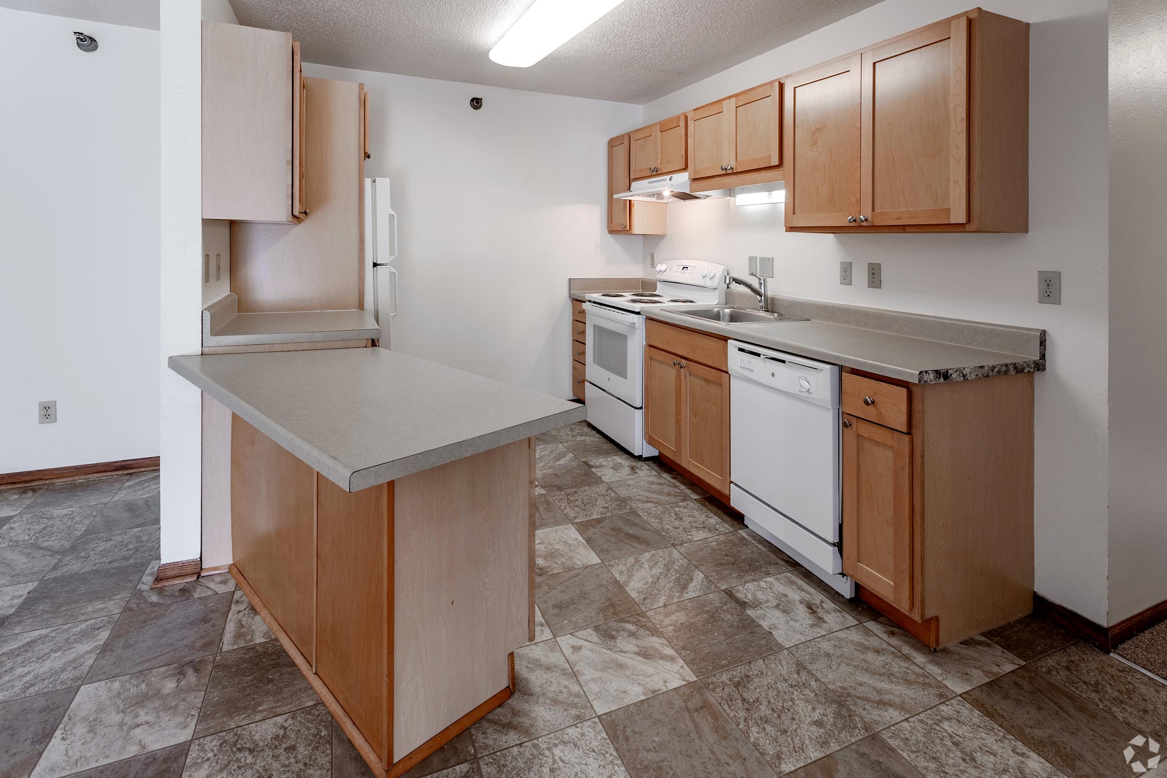 A spacious kitchen featuring beige cabinets, a countertop, and modern appliances including a refrigerator, stove, and dishwasher. The floor is tiled with a muted gray and brown pattern, creating a warm and inviting atmosphere. Natural light fills the room through a ceiling light and reflects off the surfaces.