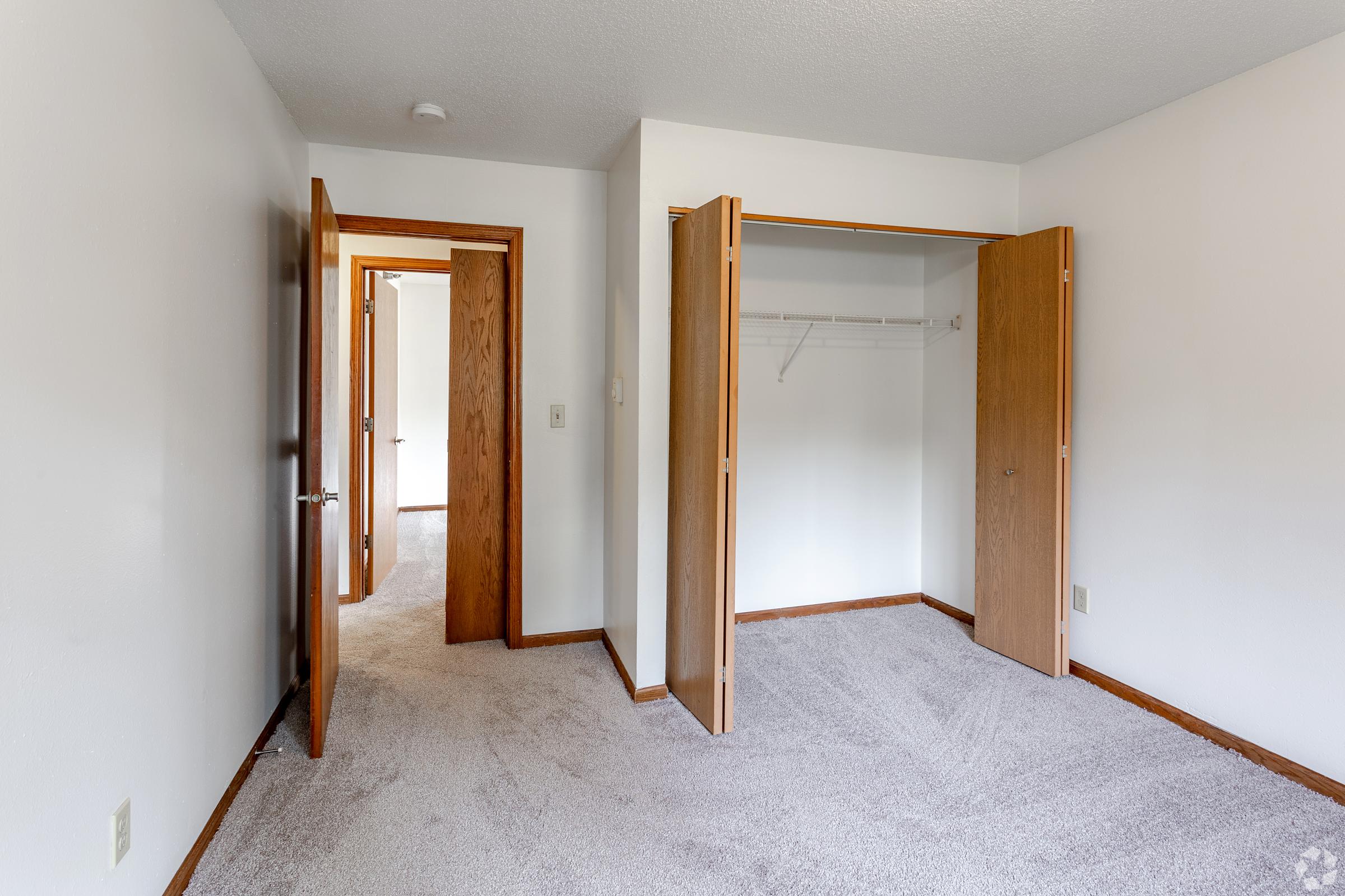 A spacious, empty room featuring light-colored walls and carpet. Two open doors lead to a closet and another room, creating an airy feeling. The room is well-lit with natural light, showcasing its potential for personalization and decor.
