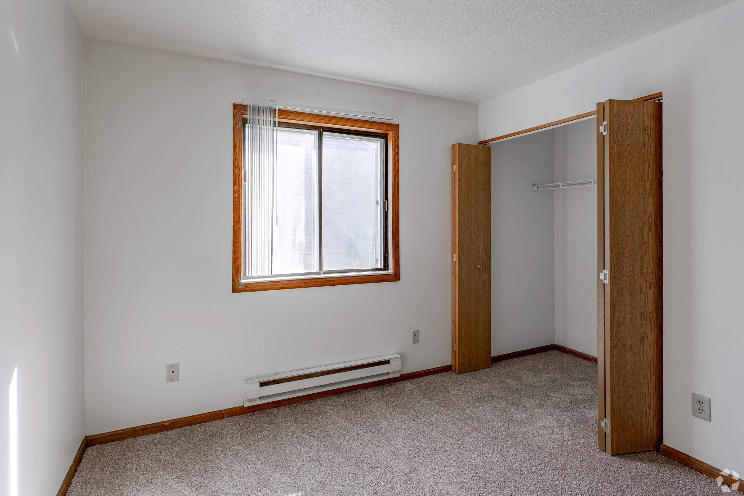 Empty bedroom with light beige carpet, white walls, and a large window with sheer curtains. There's a wooden closet with open doors on the right. The room is well-lit, showcasing a minimalistic and clean design.
