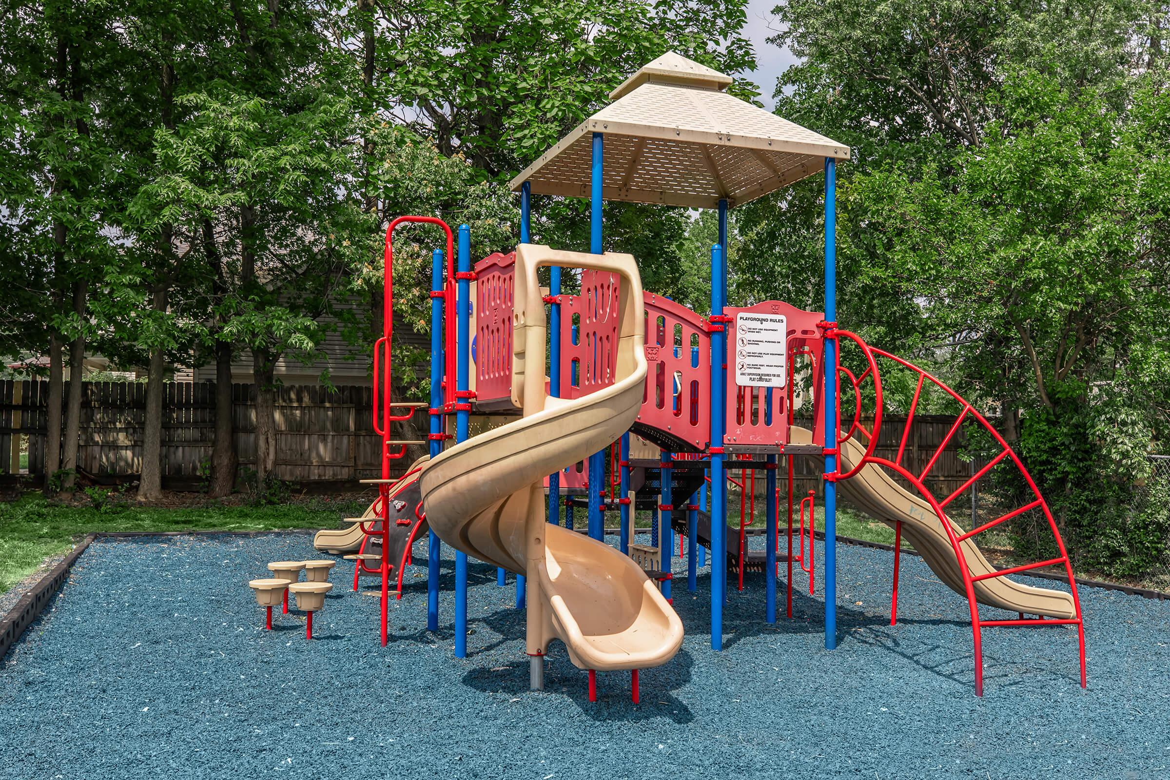 A colorful playground featuring a tall play structure with slides, climbing walls, and an enclosed area, set on a bed of blue safety surface. Surrounded by green trees and a wooden fence, the playground offers a fun and safe space for children to play and explore.