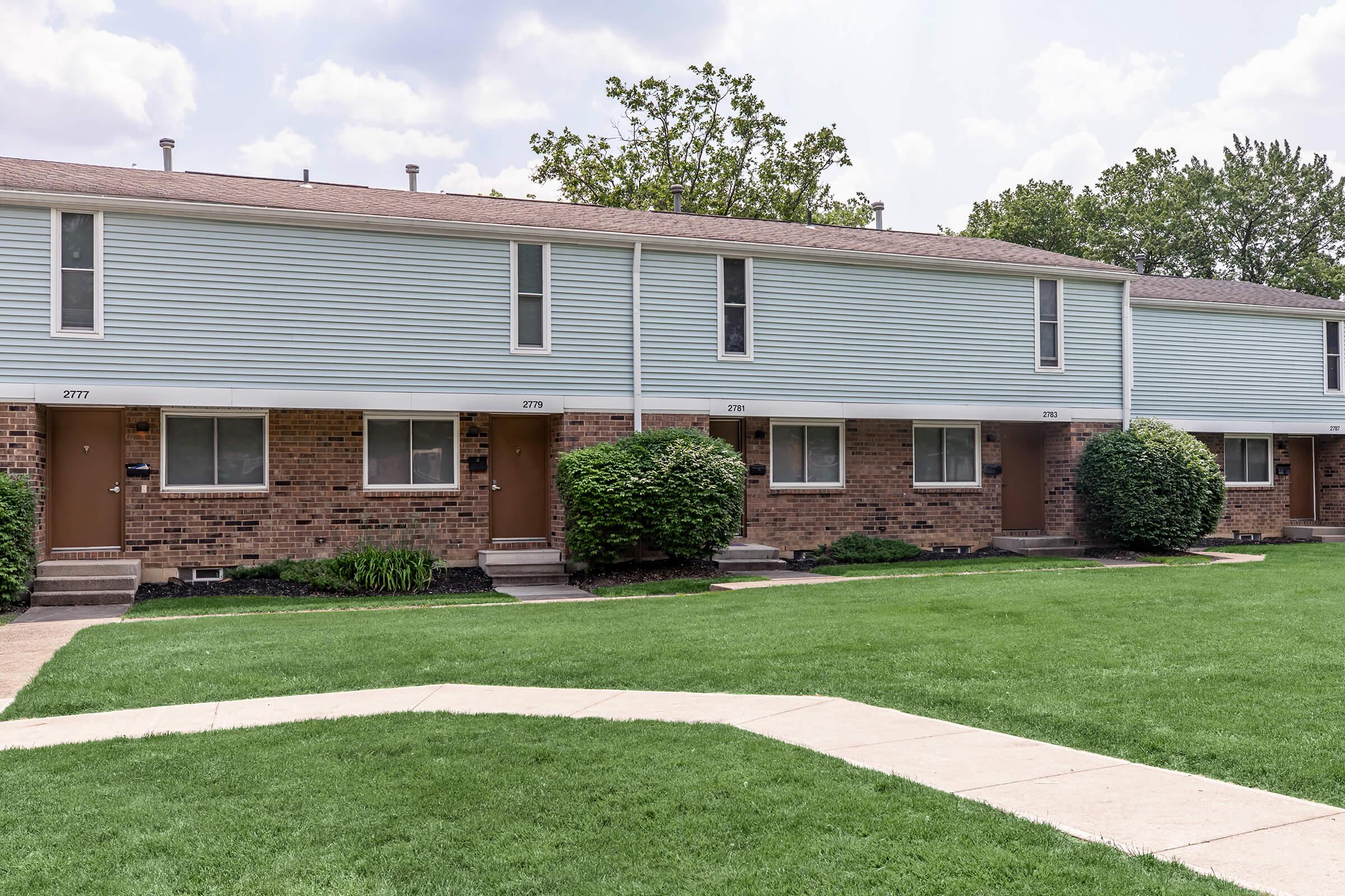 A row of townhouses with light blue siding and brick accents. Each unit has a wooden door and a small front porch. A well-manicured lawn is in front, with a concrete walkway leading to the doors. The sky is partly cloudy, and trees are visible in the background.