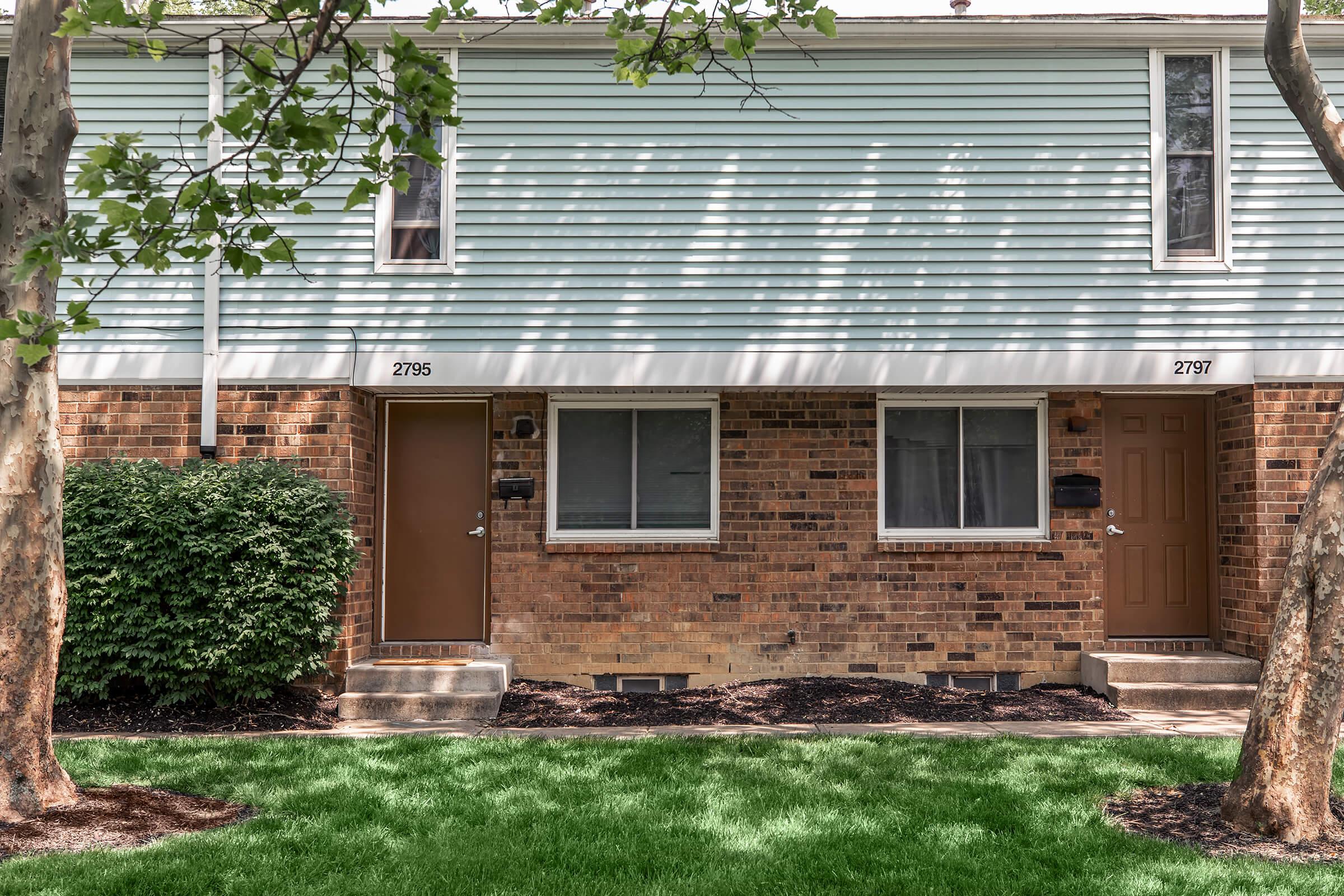 Two side-by-side townhouses with light blue siding and brick lower sections. Each unit has a separate entrance with a brown door and mailbox. The front yard features well-manicured green grass and two trees providing shade. The building numbers 2796 and 2797 are displayed above the doors.