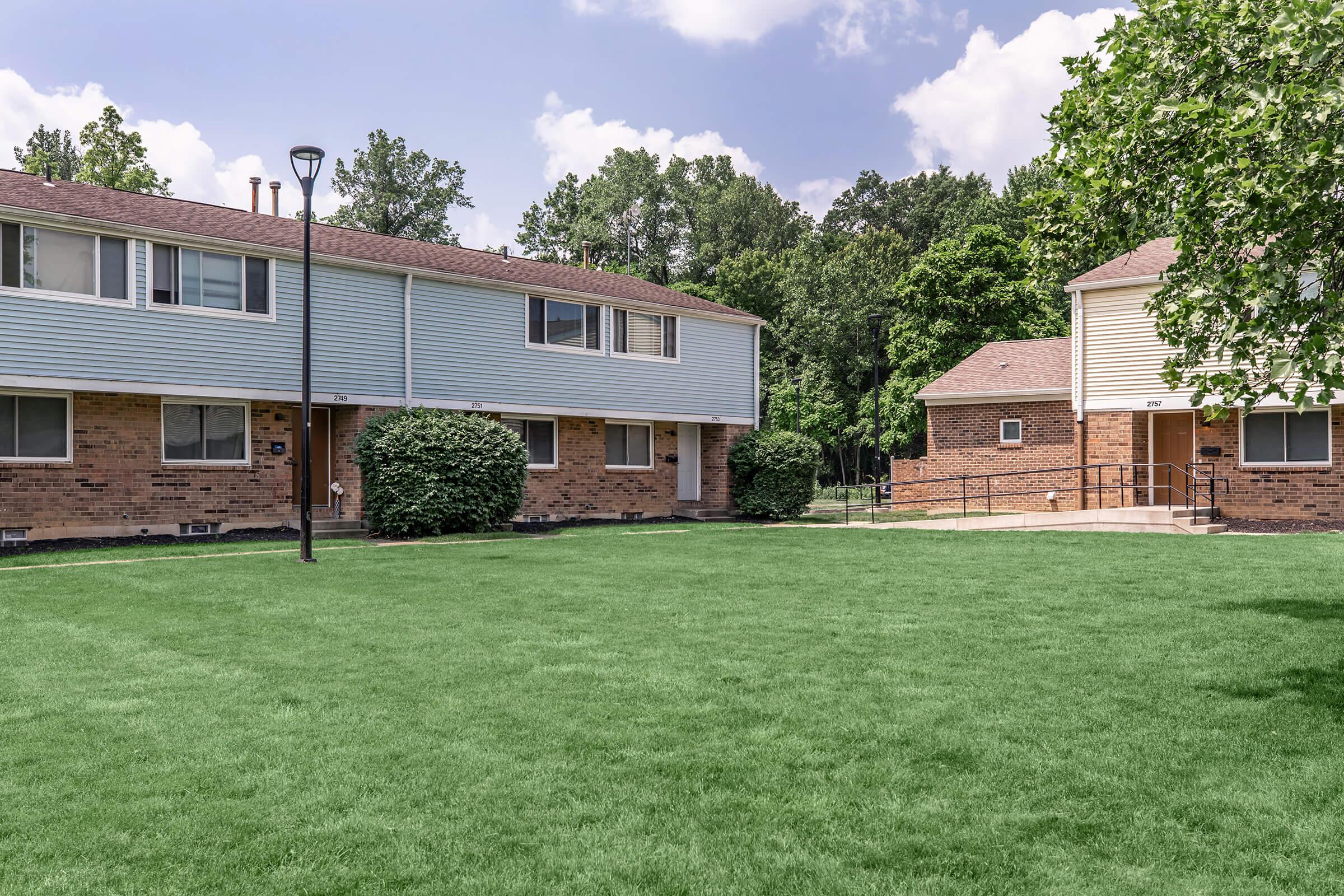 A well-maintained residential complex featuring two-story brick and siding units surrounded by lush green grass and shrubbery. A light pole stands in the foreground, and a pathway leads to a nearby building, with trees providing shade in the background under a partly cloudy sky.