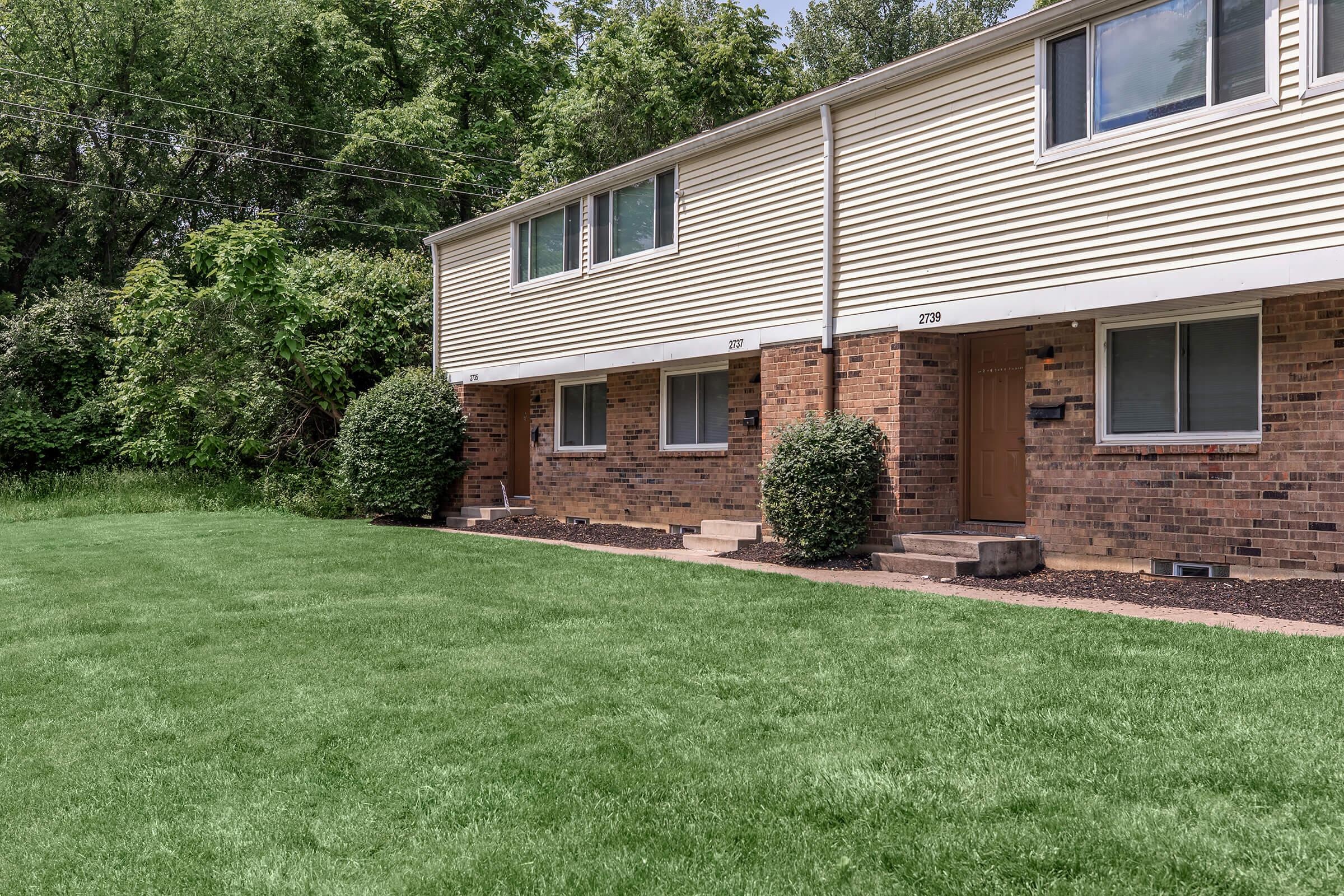 Row of two-story townhouses with brick and beige siding, featuring green lawns in front. Each unit has a small porch and a neatly trimmed bush beside the entrance. Surrounding trees and greenery provide a natural backdrop, suggesting a quiet residential area.