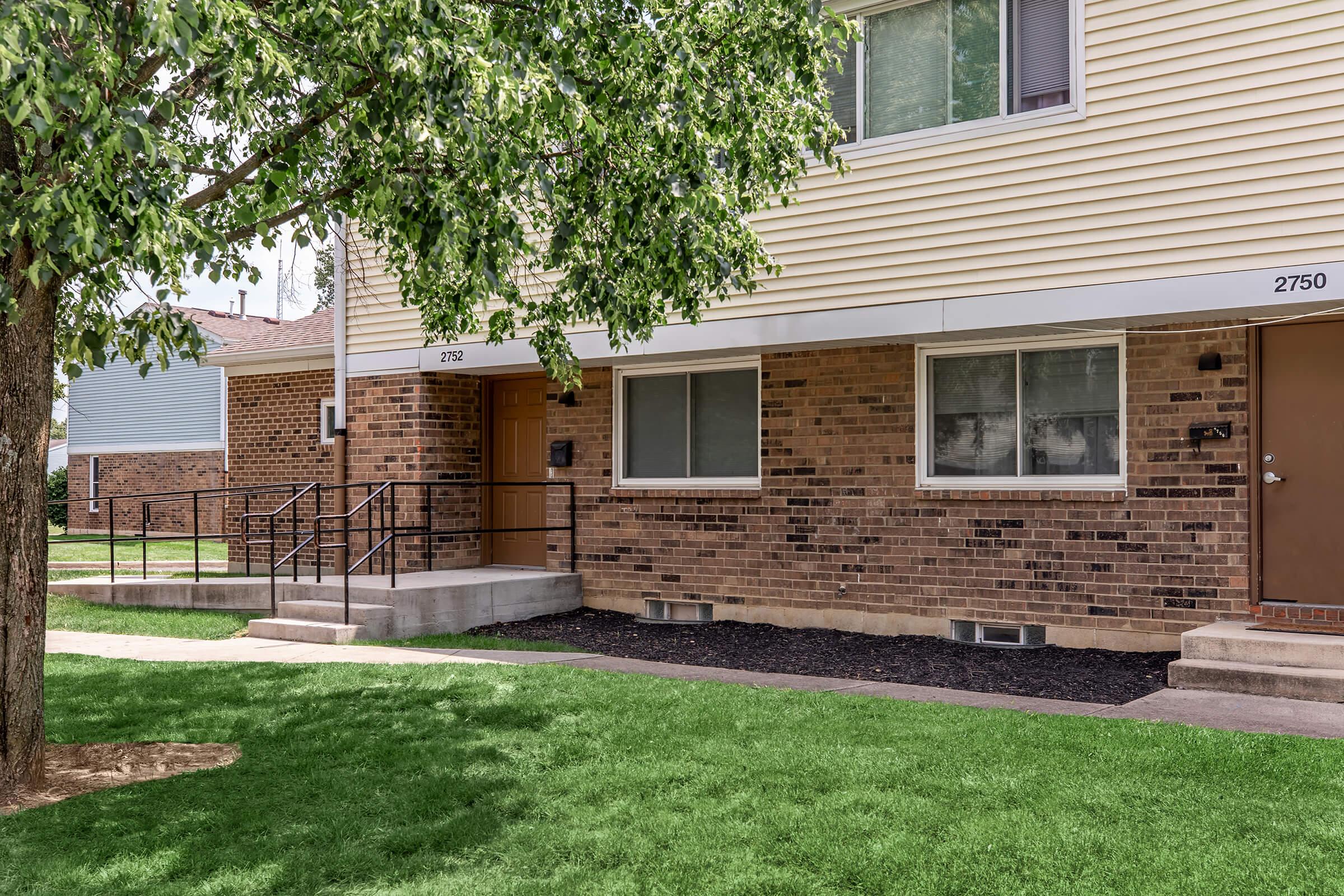 Two residential buildings with brick exteriors and light-colored siding. The foreground features a well-manicured green lawn with a small tree. The first building has accessible ramp access, while the neighboring building has a simple door entrance.