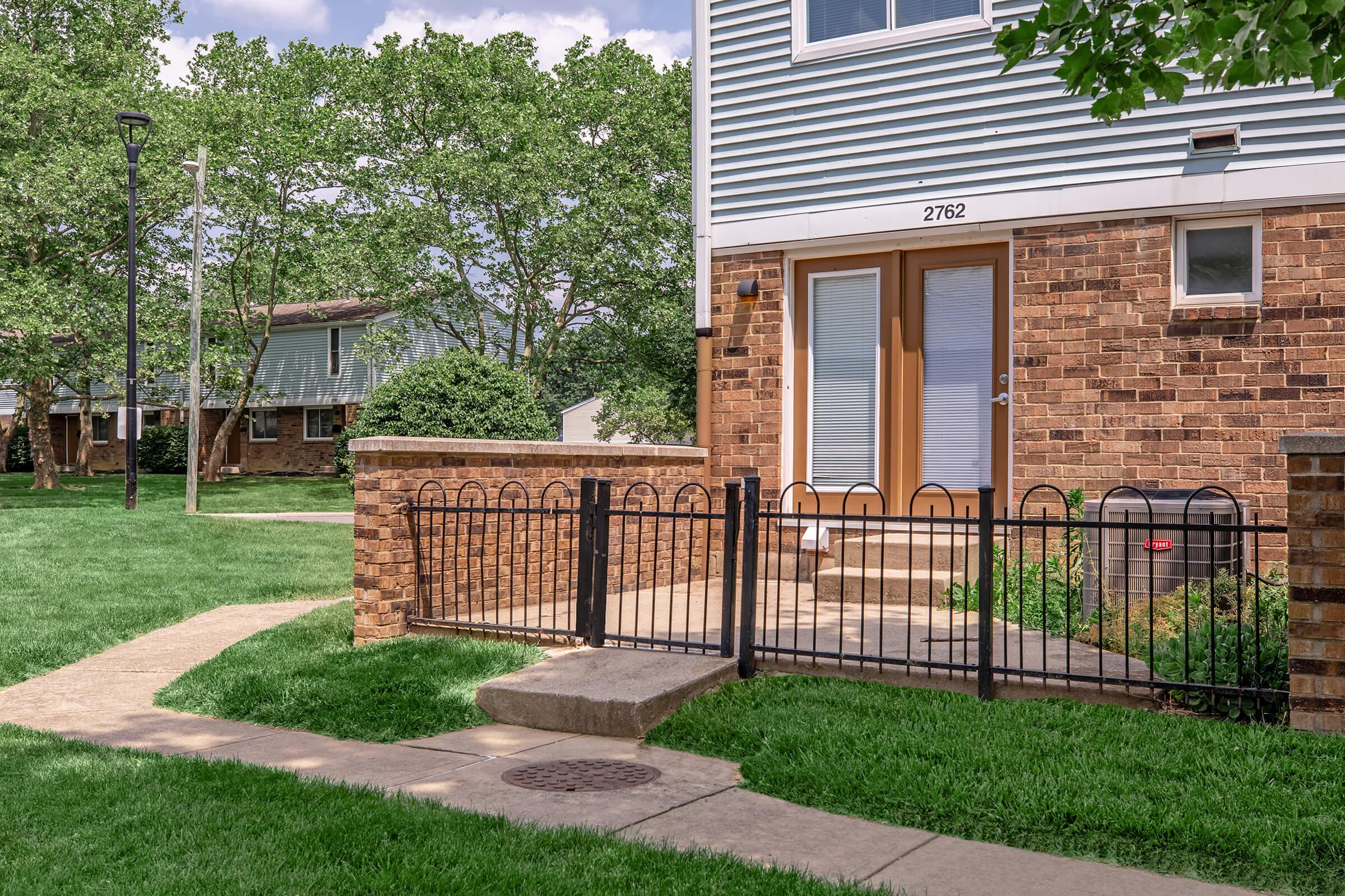 A corner view of a brown brick building with a light blue siding, featuring a fenced entrance and paved walkway. The surrounding area is grassy, with trees and a sidewalk visible. The building has large windows and a front door with a small porch.