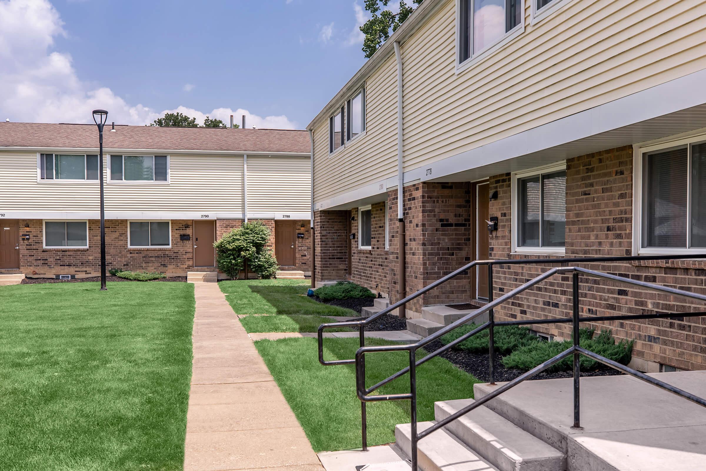Two residential buildings with a mix of brick and siding exteriors. A well-maintained green lawn is visible in front of the units, along with a concrete pathway leading up to the entrances. Each unit features a small porch area with steps, and there are lamp posts lining the pathway.
