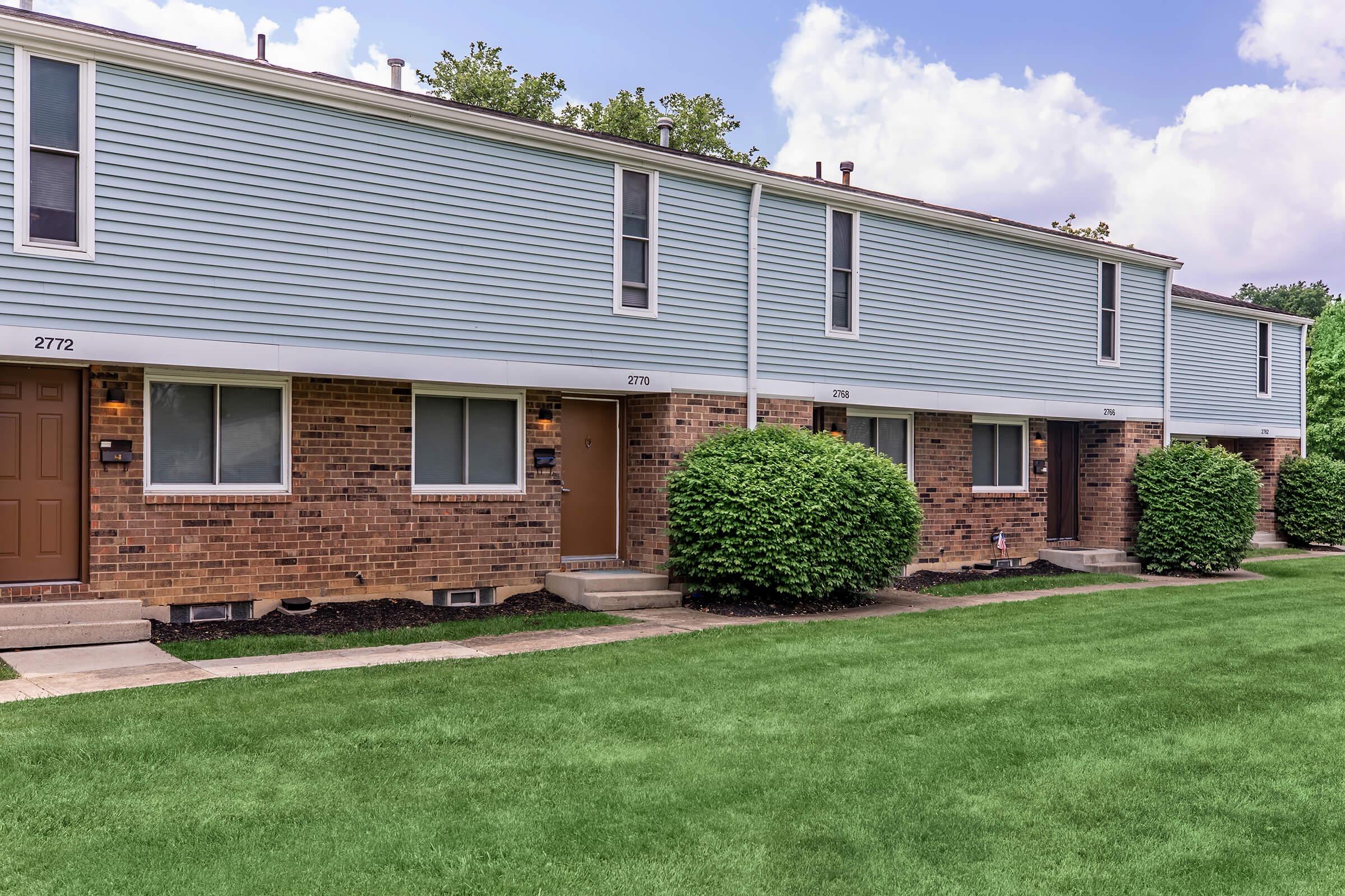 Row of townhouses with a mix of blue siding and brick facades. Each townhouse has a front door with a small porch and windows. The grassy area in front is well-maintained, with hedges lining the walkway. The sky is partly cloudy, creating a pleasant neighborhood scene.