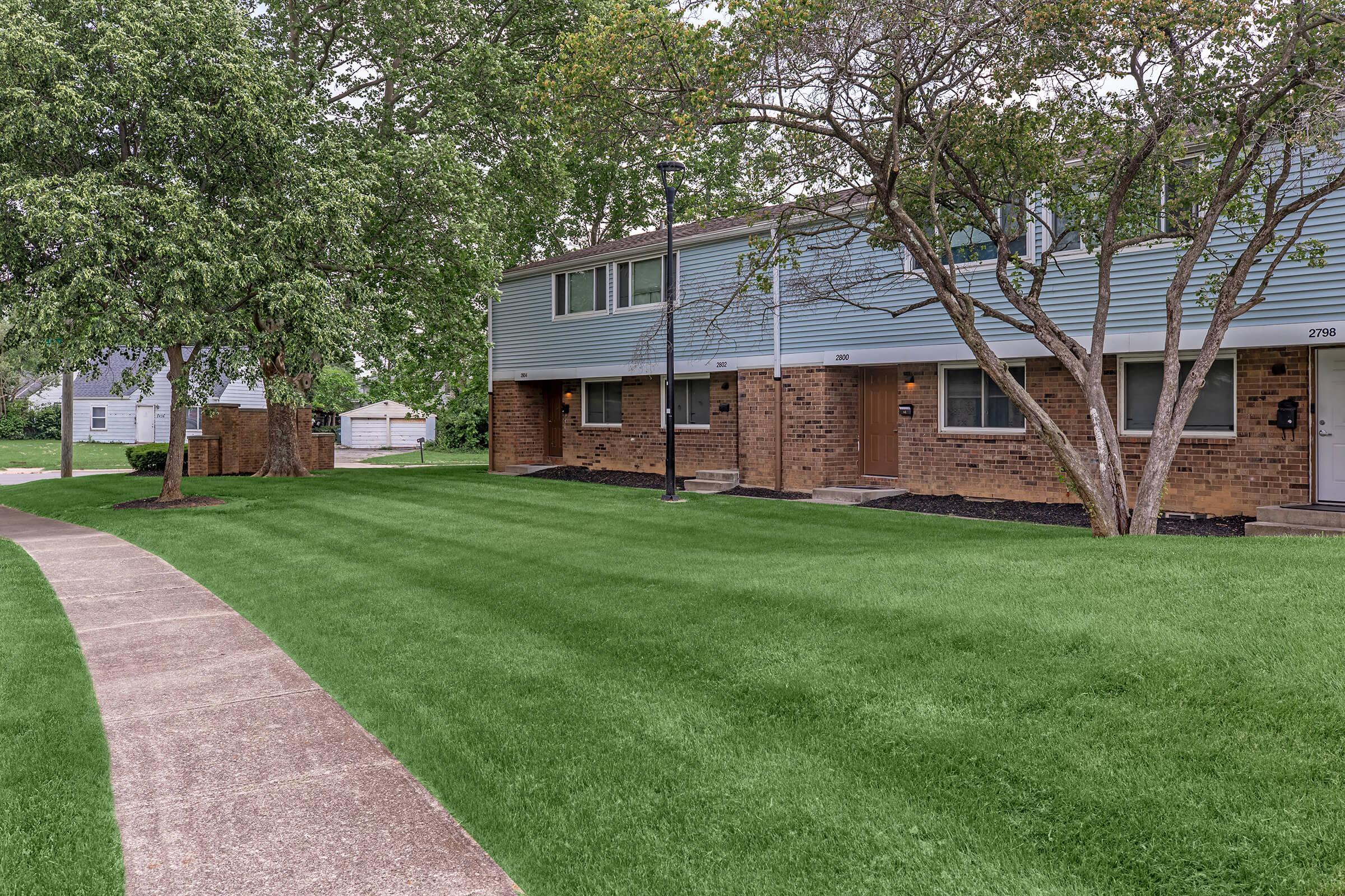 A well-maintained residential area featuring a two-story apartment building with a grassy lawn, trees, and a paved walkway. The building has brick and siding exteriors, with windows visible on each floor. Pathways lead to the entrance, and there are nearby parking spaces and landscaping elements.