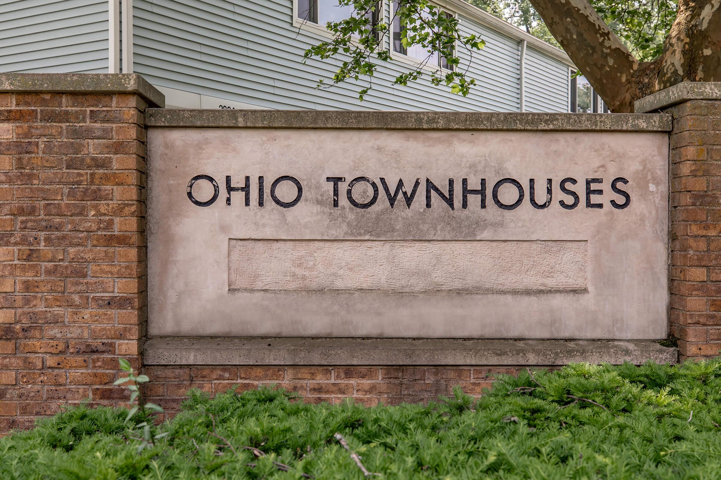 Sign for "Ohio Townhouses" made of brick and concrete, featuring bold, engraved lettering on a light-colored surface. Surrounded by greenery and set against a residential building in the background.