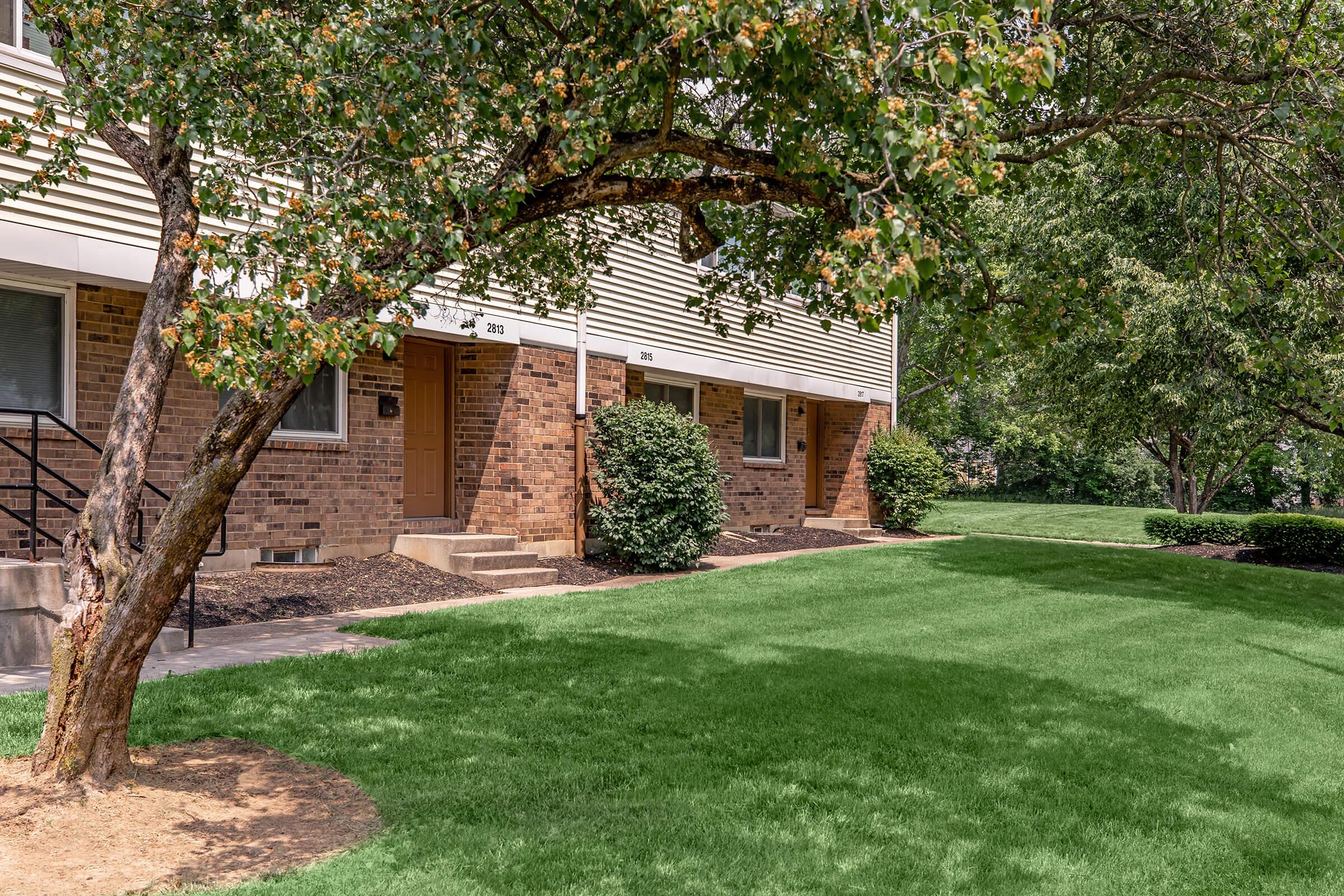 A row of brick apartment buildings with tan siding, featuring front doors numbered 201 and 202. A grassy lawn stretches in front, with a small tree on the left side and well-trimmed bushes nearby. The scene is sunny and well-maintained, showcasing a peaceful residential environment.
