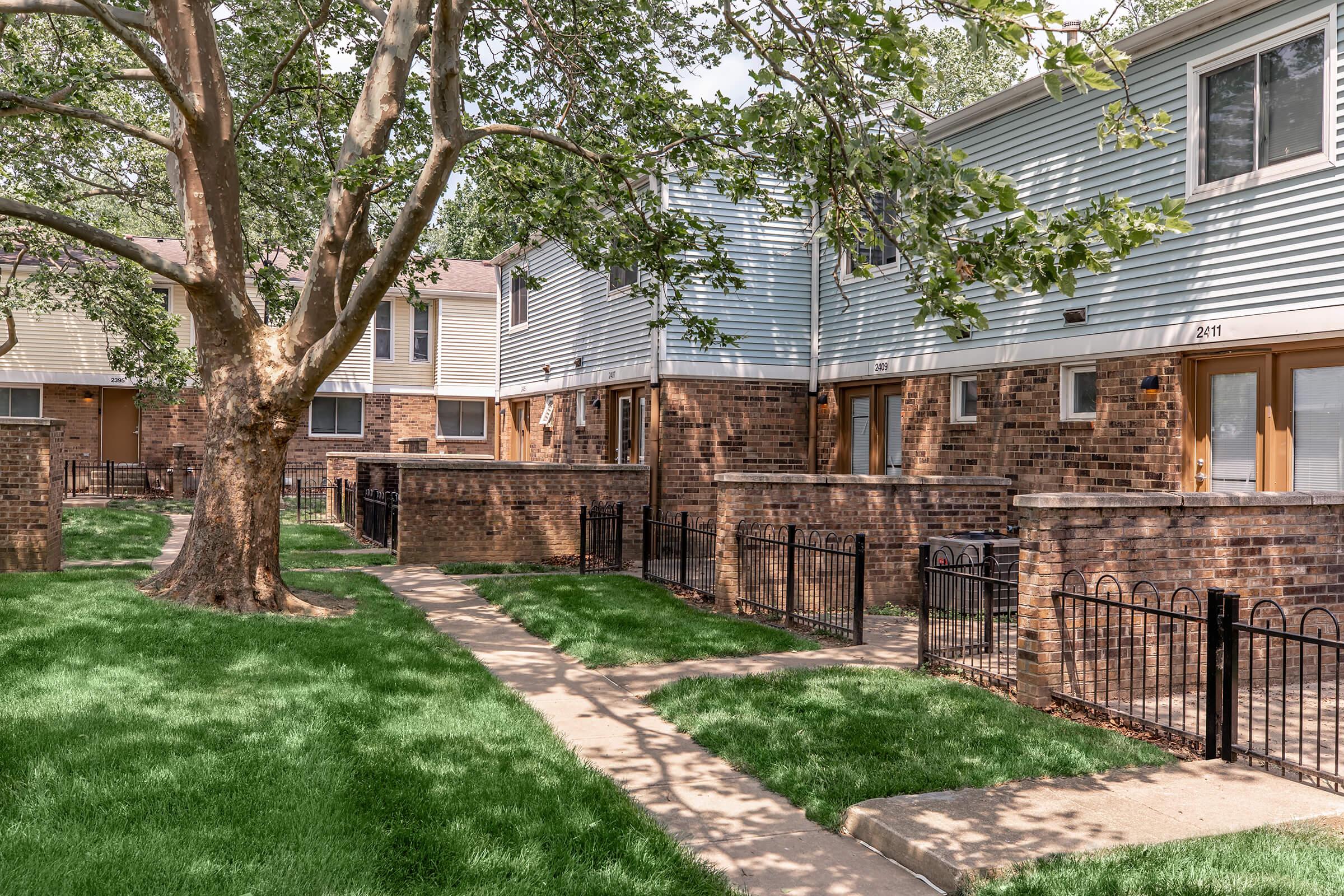 View of a residential area featuring two-story apartment buildings made of brick and siding. A large tree stands in the foreground, surrounded by green grass and neatly trimmed pathways. The buildings have fenced patios and are set in a suburban neighborhood with clear blue skies.