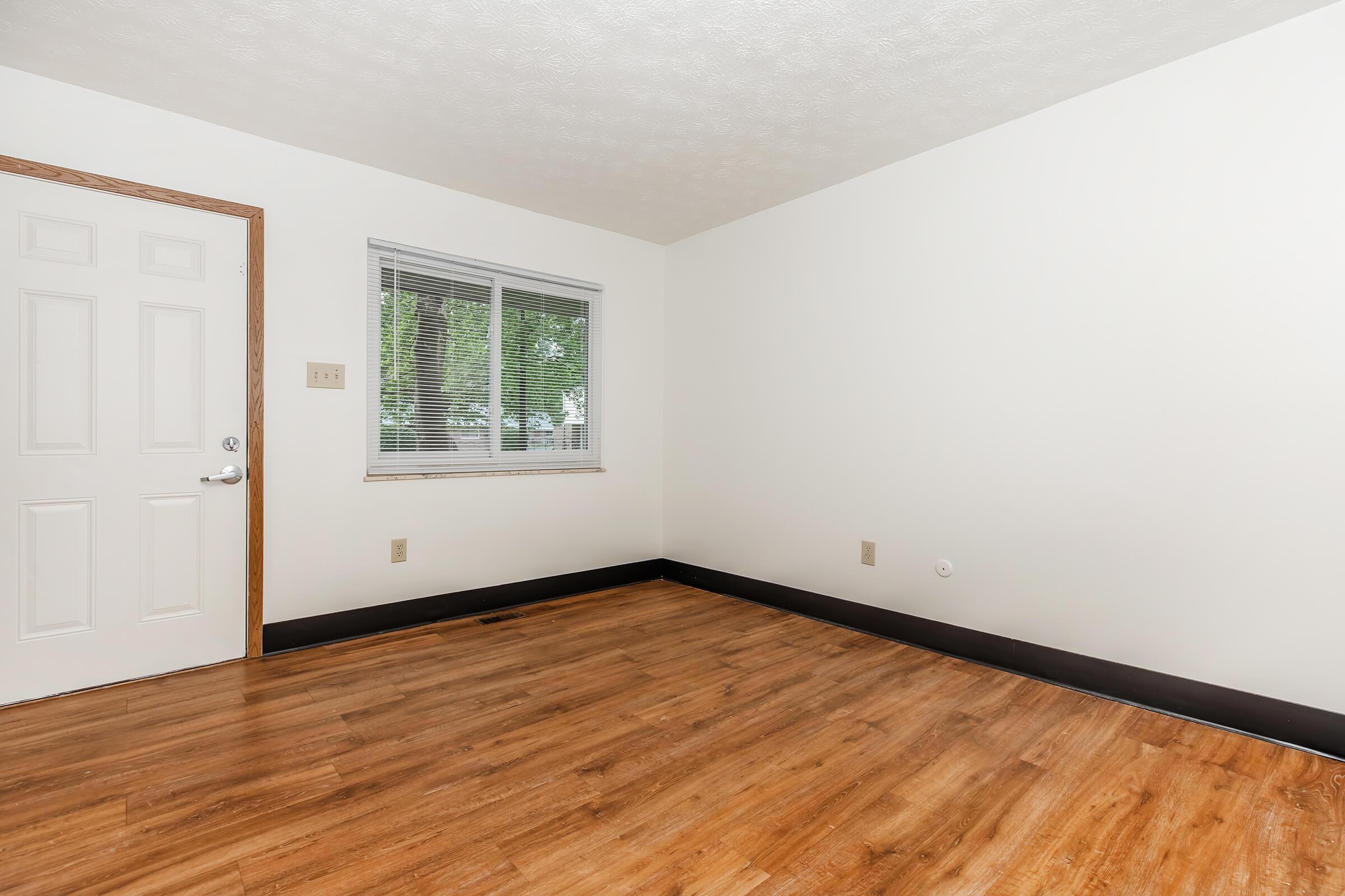 A spacious, empty room with light-colored walls and a wooden floor. There is a window on one side allowing natural light, and a white door on the adjacent wall. The room appears clean and well-maintained, suitable for various uses such as a bedroom or office.