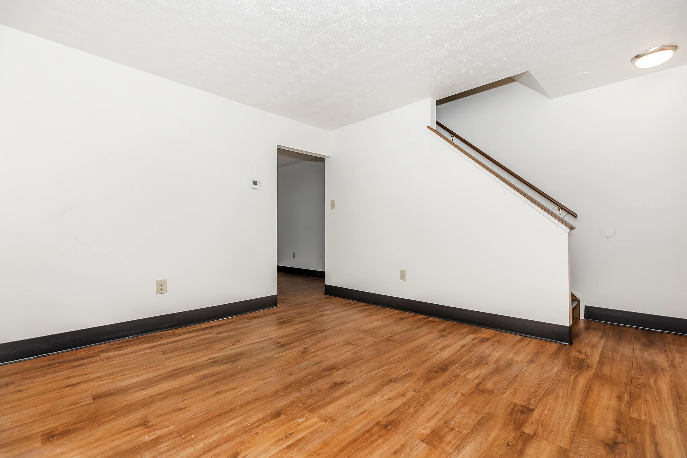 Interior of a spacious room with light-colored walls and wooden flooring. A staircase is visible in the background, and there is a door leading to another room. The space is well-lit and uncluttered, providing a minimalist and open feel.