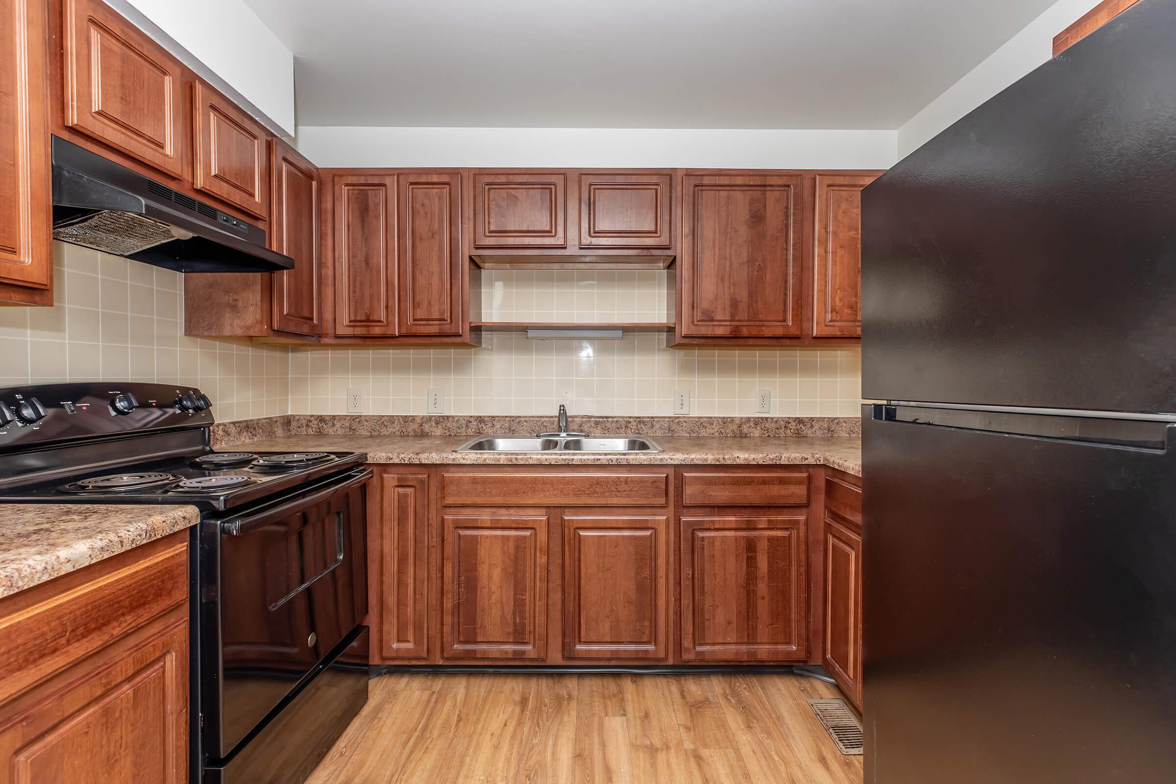 A modern kitchen featuring wooden cabinets, black appliances including an oven, stove, and refrigerator. The countertop is made of a light-colored material, and a double sink is positioned under a wall-mounted cabinet. The flooring is a light wood finish, creating a warm and inviting atmosphere.
