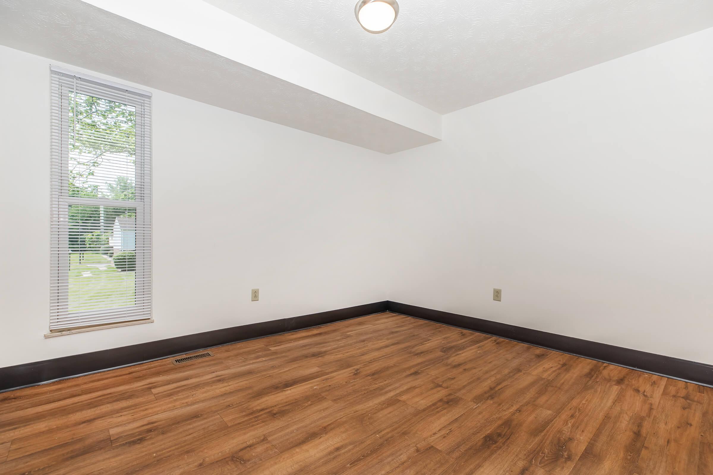 Empty room featuring light-colored walls, a single window with blinds allowing natural light, and wooden flooring. A ceiling light fixture is visible, and the room appears spacious with no furniture. The view outside the window shows green grass and trees.