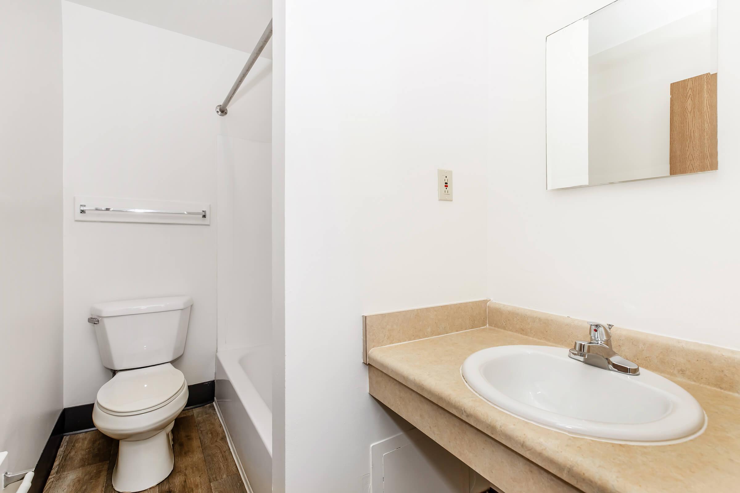 A small bathroom with a bathtub and shower combination on the left, a white toilet, and a light-colored countertop with a sink. A mirror is mounted above the sink, and the walls are painted white, giving the space a clean and simple look. The flooring appears to be a wood-like laminate.