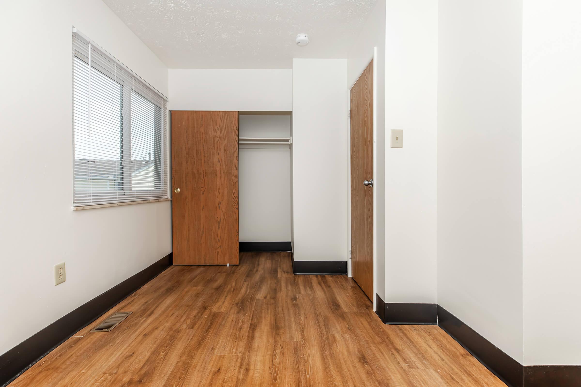 A vacant, well-lit room featuring light wooden flooring. There is a window with blinds on the left, an open wooden closet door on the left side, and a closed wooden door on the right. The walls are painted white, creating a clean and spacious atmosphere.