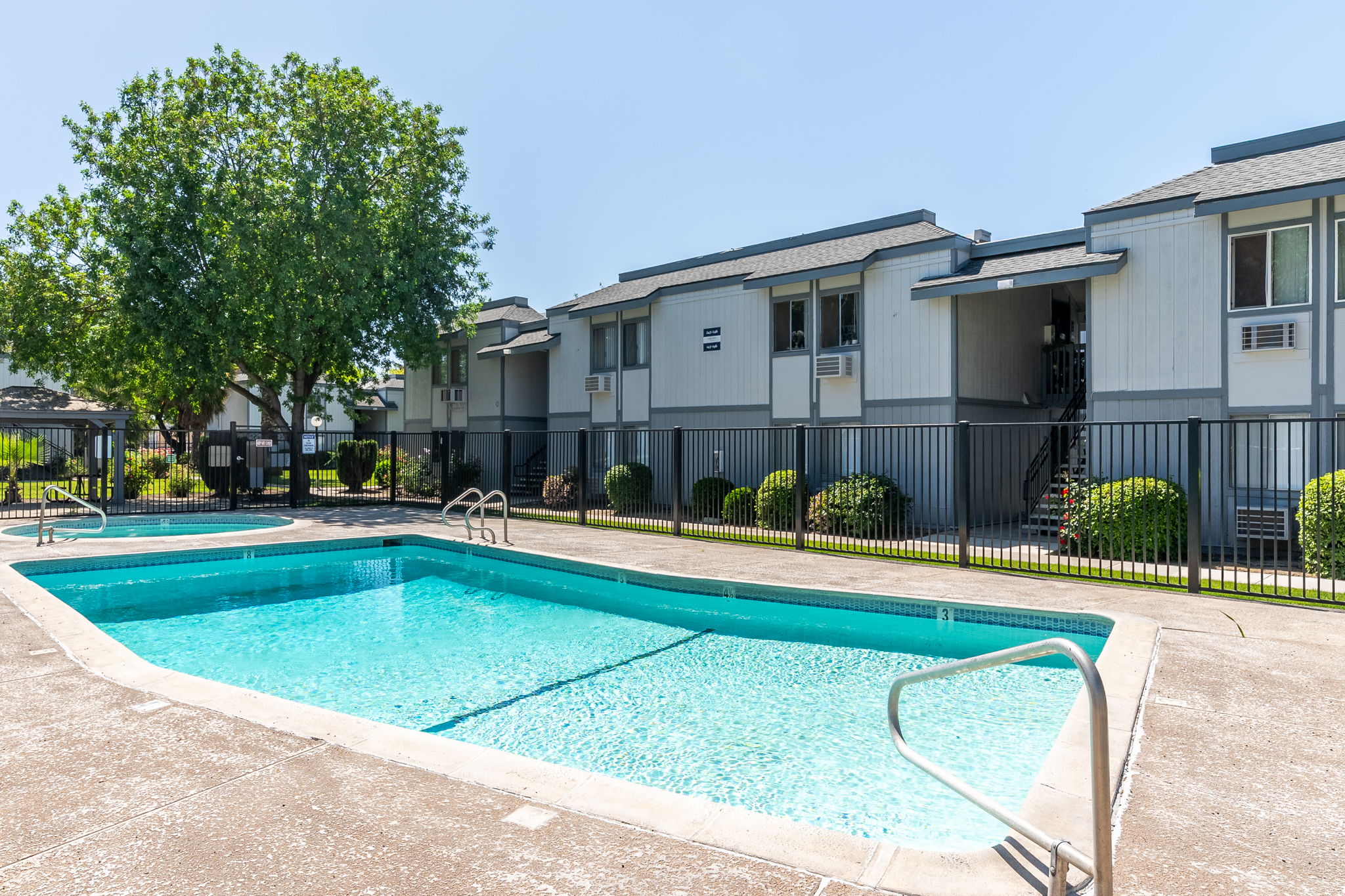 A sparkling blue swimming pool surrounded by a black fence, with a shaded area featuring a large tree. In the background, there are two-story apartment buildings with balconies. The scene is bright and sunny, capturing a relaxing outdoor space ideal for leisure and recreation.