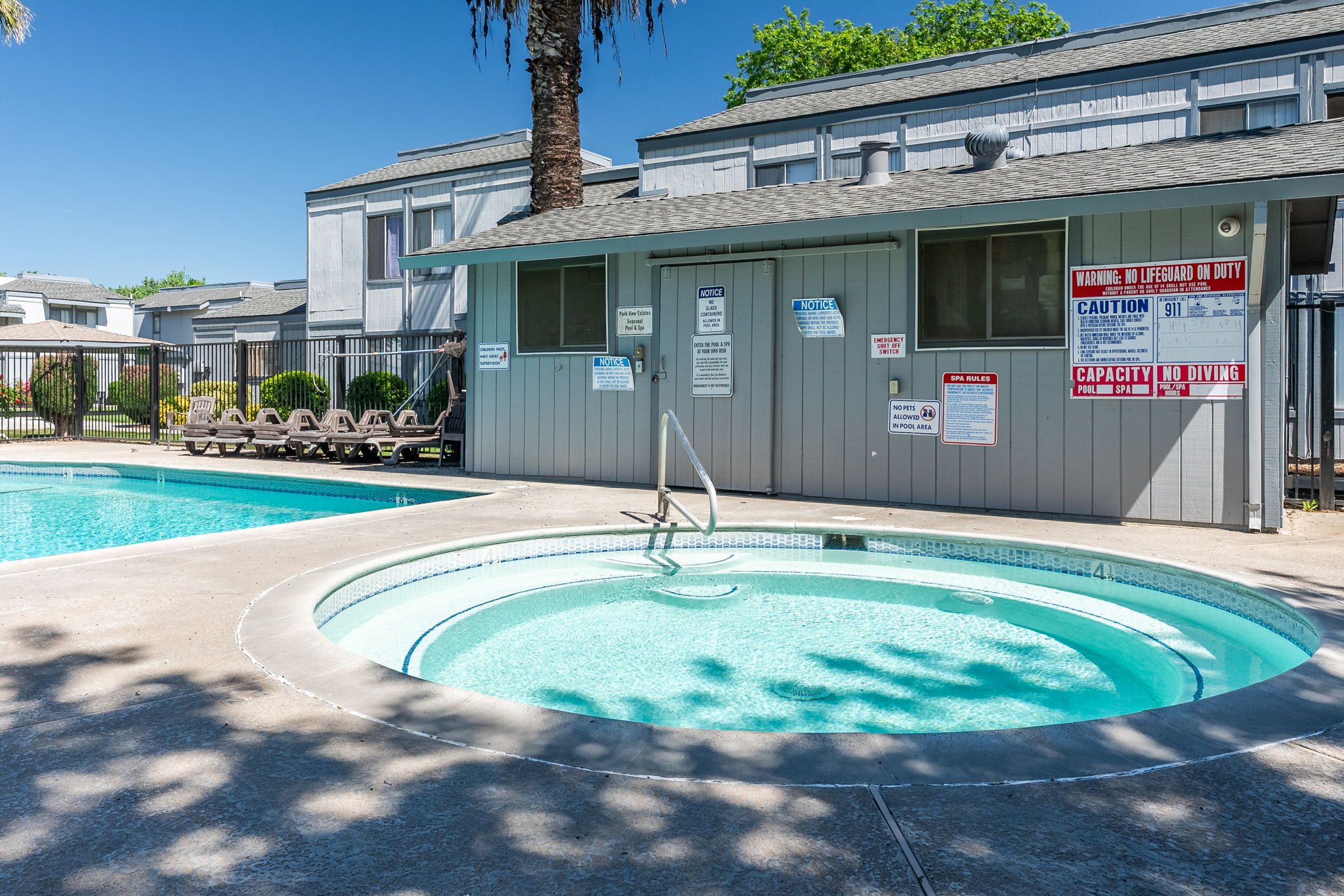 A sunny outdoor area featuring a swimming pool and a hot tub. The pool is surrounded by lounge chairs, with a fence in the background. Nearby, there is a building with multiple signs indicating safety guidelines and pool rules. Lush green trees add to the overall ambiance of the scene.