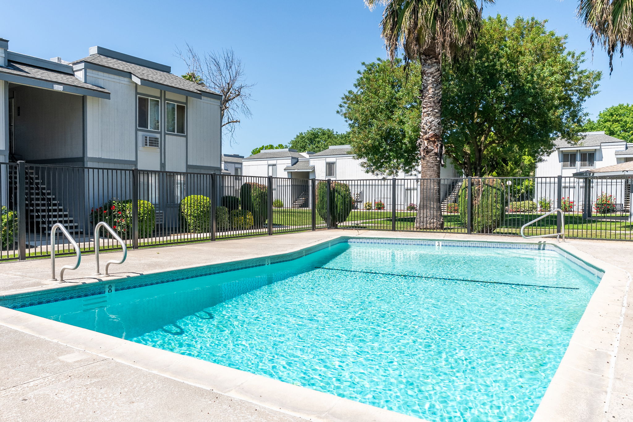 A clear swimming pool surrounded by a fence, with palm trees and well-manicured lawns in the background. Apartment buildings are visible, indicating a residential complex. The sky is bright blue, creating a sunny and inviting atmosphere.