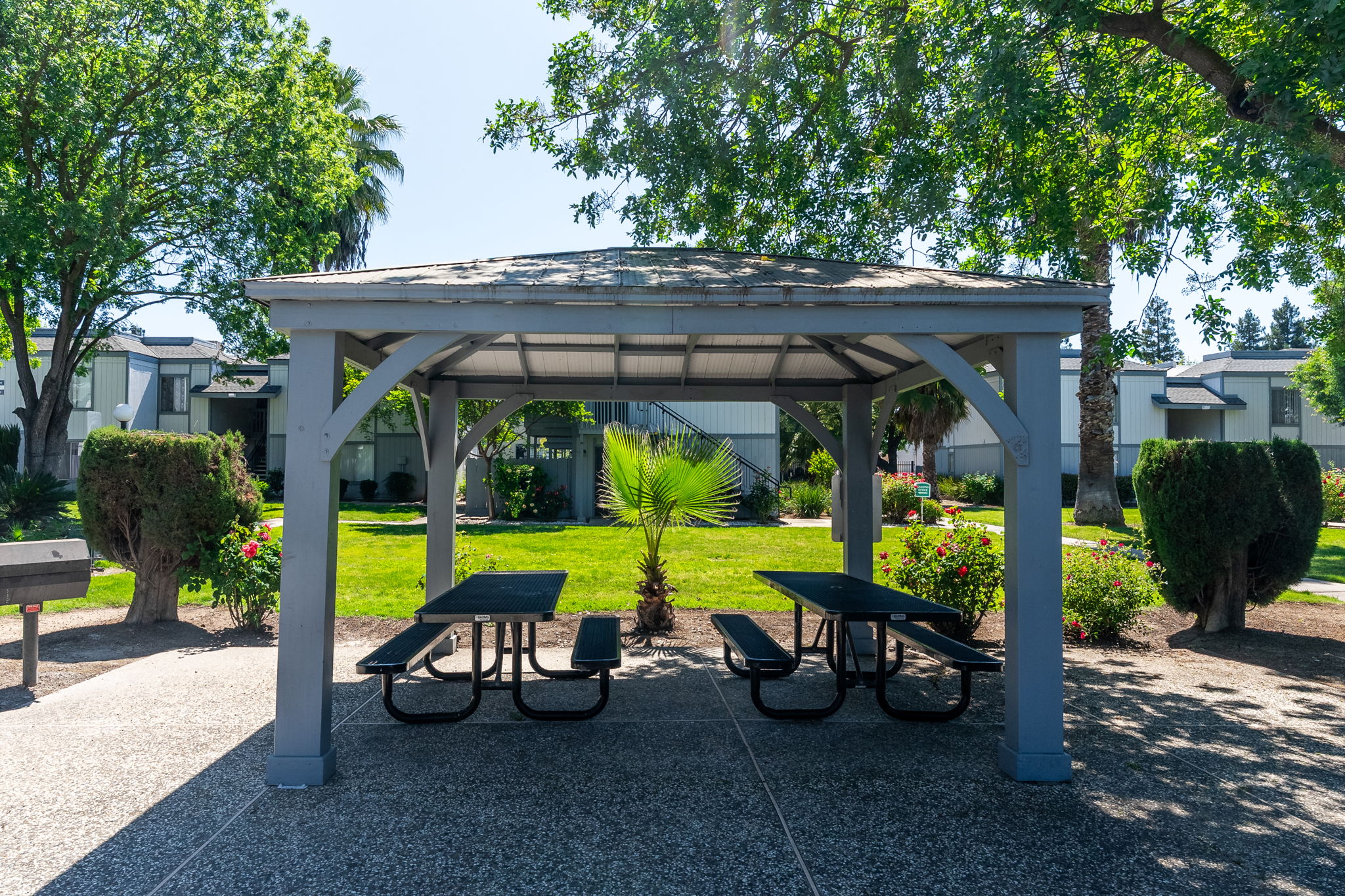 A shaded gazebo with several picnic tables underneath, surrounded by well-maintained greenery, including trees and shrubs. The area appears inviting and ideal for outdoor gatherings or relaxation. Sunlight filters through the leaves, creating a pleasant atmosphere.