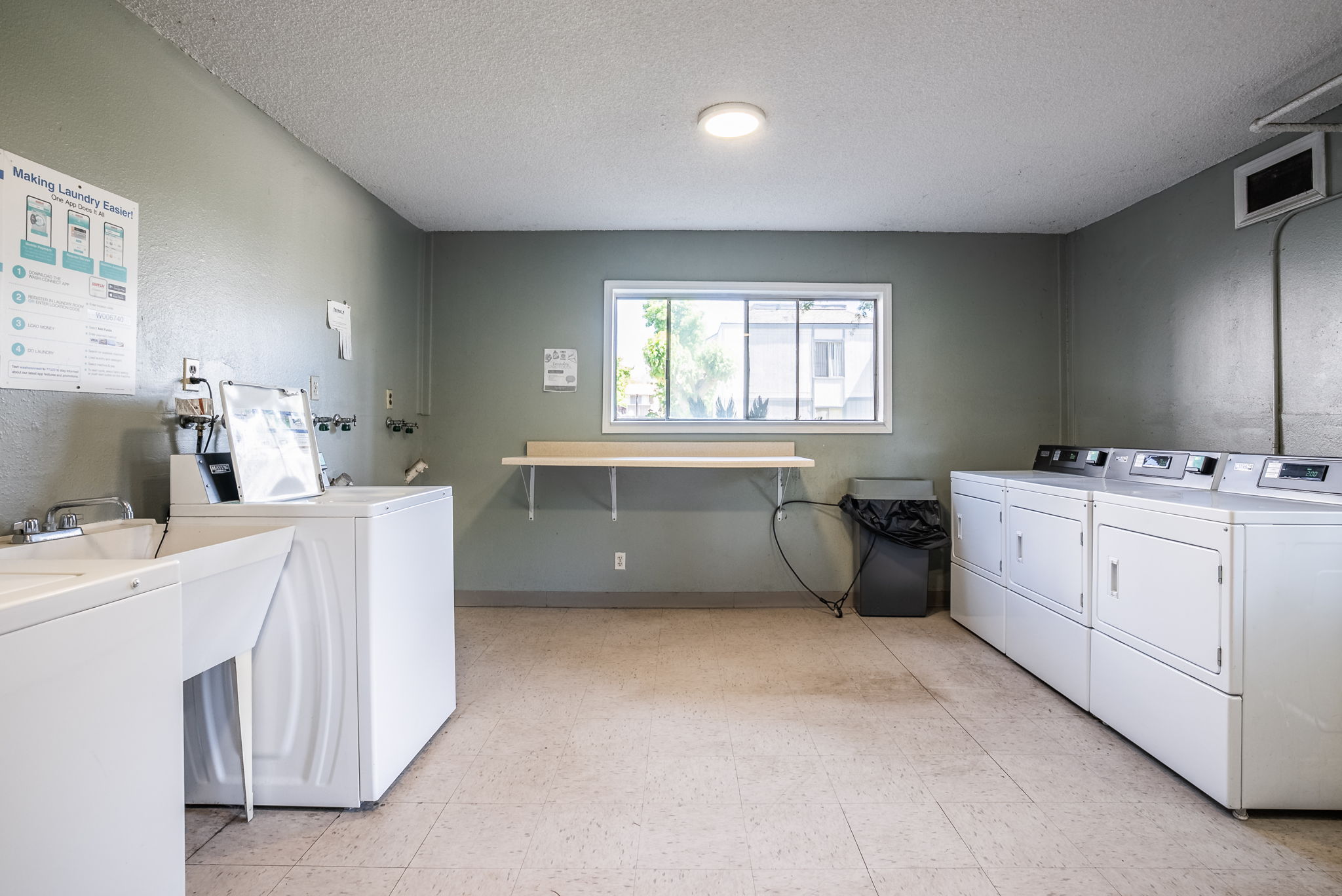 A clean laundry room featuring several washing machines and dryers against green walls. A utility sink is visible, along with a shelf by a window. There’s a waste bin in the corner, creating a functional space for washing and drying clothes.