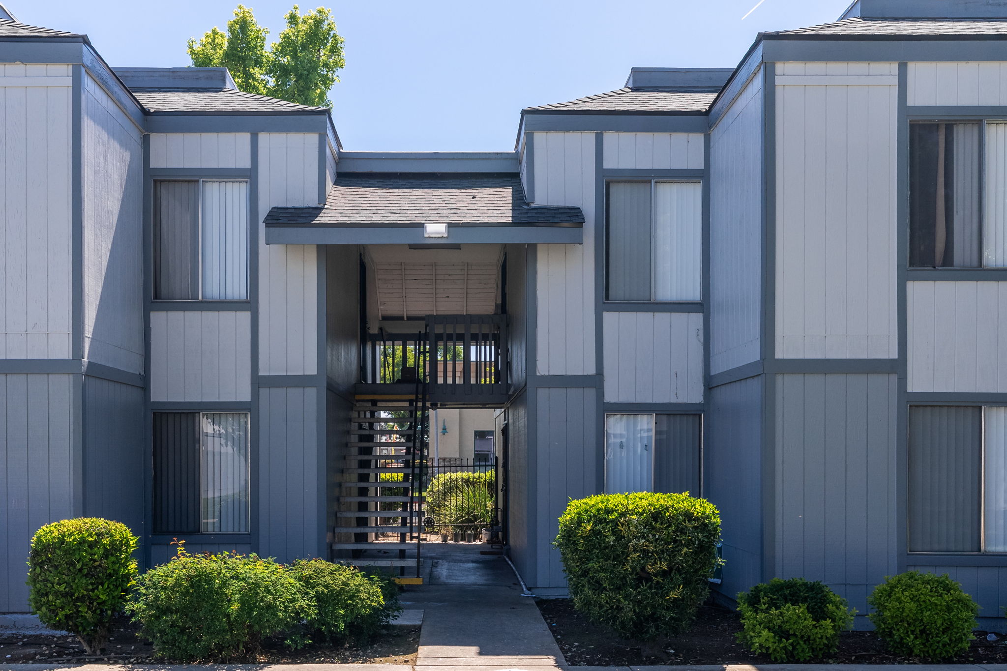 A view of a modern apartment complex featuring two gray buildings with a central staircase. The entrance is framed by greenery, including neatly trimmed bushes. Bright sunlight illuminates the scene, giving it a welcoming atmosphere. The sky is clear with a hint of blue.