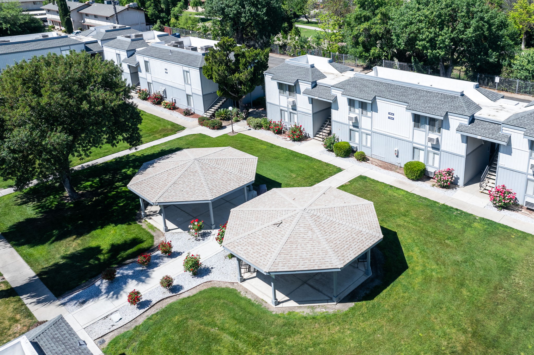 Aerial view of a housing complex featuring multiple single-story buildings with gray roofs. Two octagonal gazebos are located on a well-maintained lawn, surrounded by flower gardens. The area is tree-lined, and pathways connect the buildings and gazebos, creating a serene residential environment.