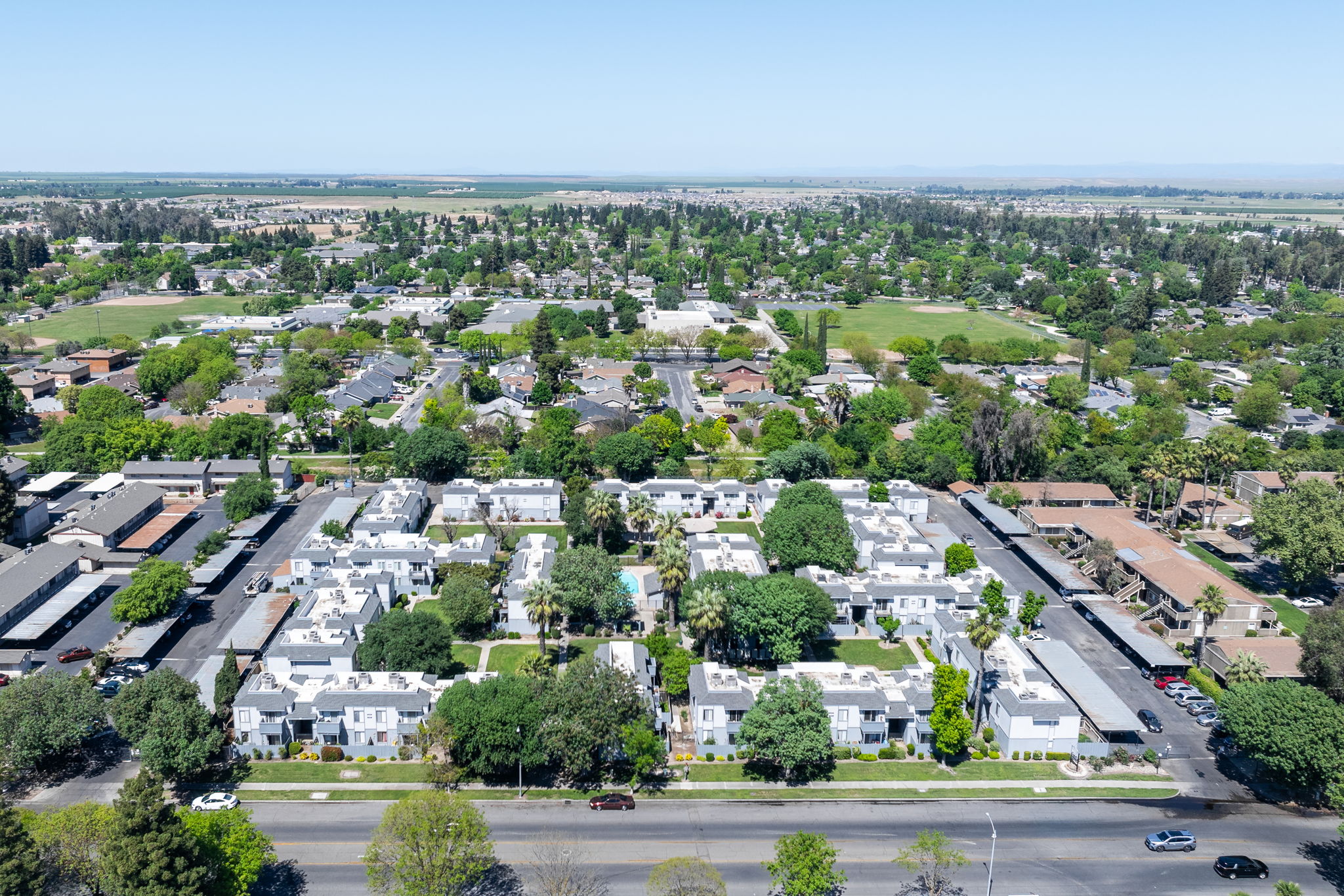 Aerial view of a residential neighborhood featuring a cluster of white apartment buildings surrounded by green trees and lawns. The area is bordered by roads, with additional houses and open spaces visible in the distance, showcasing a mix of urban and suburban landscapes.